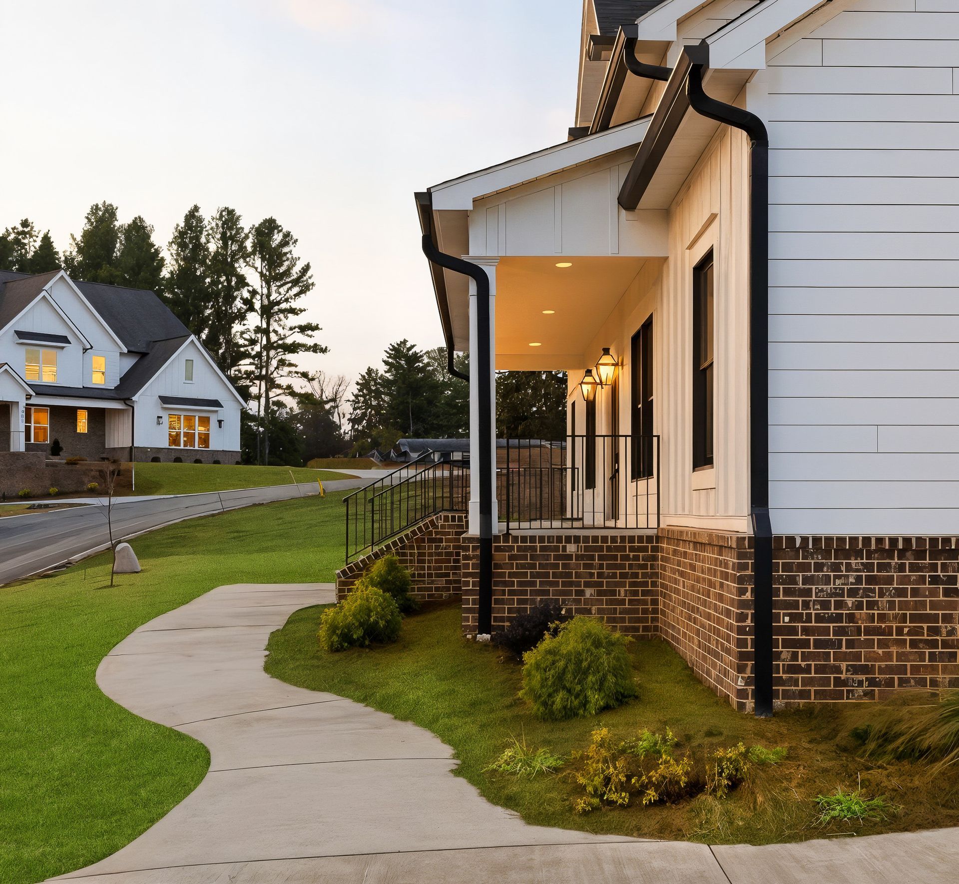 Curved sidewalk leading to a brick-and-white suburban house with a lit front porch at dusk