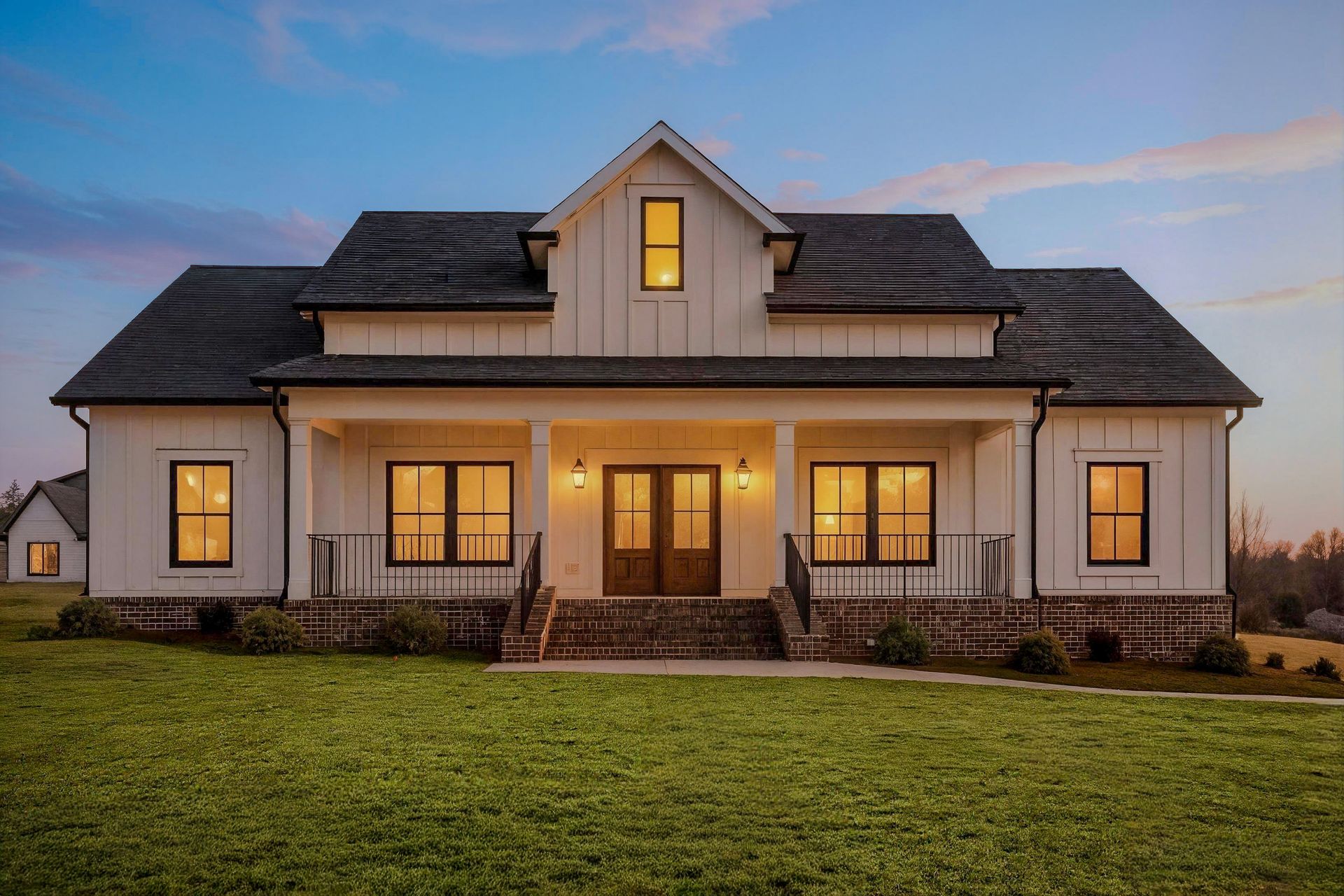 Two-story house with lit windows at dusk and a front porch, set on a grassy lot
