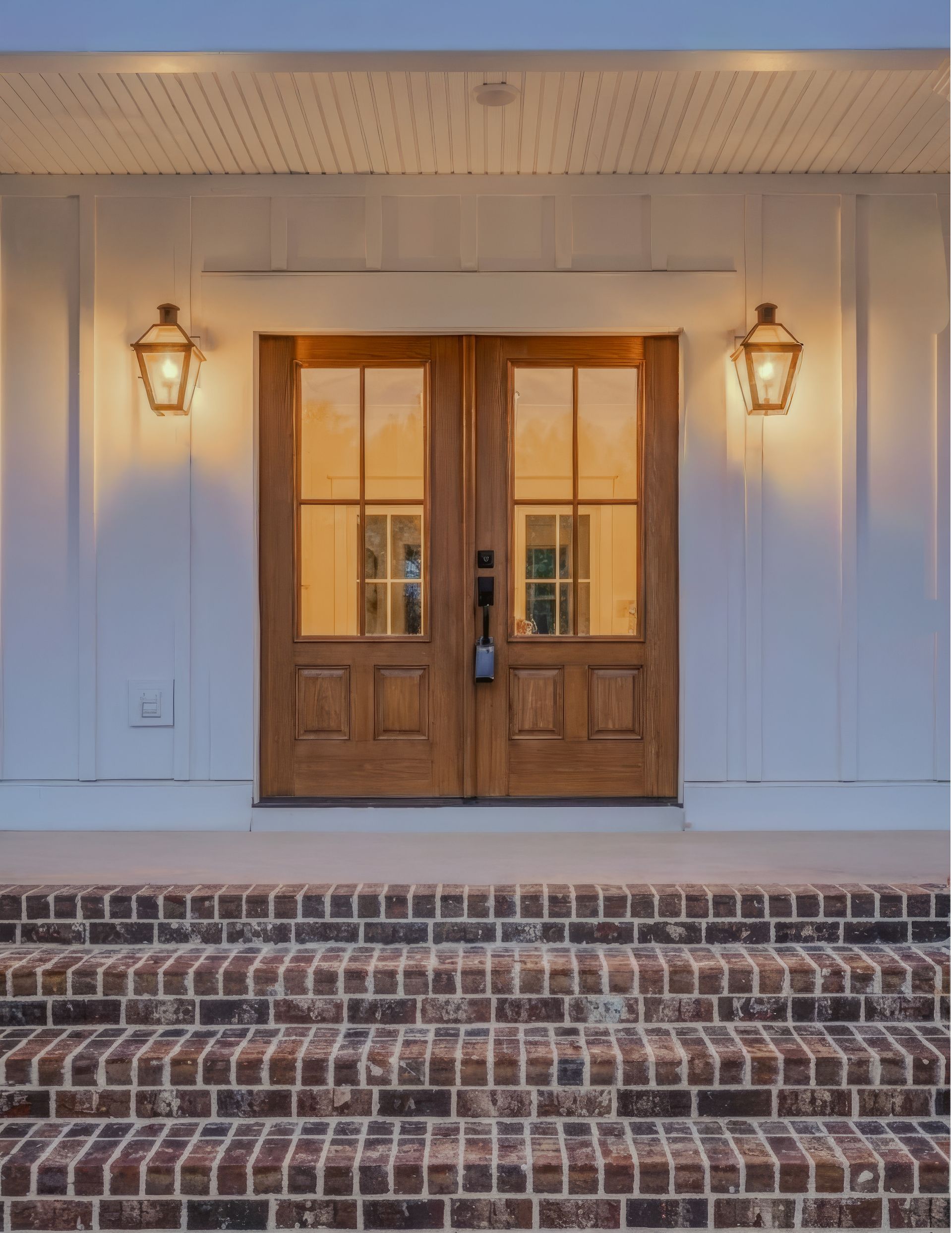 Front entrance with wooden double doors, lanterns, and brick steps on a white porch