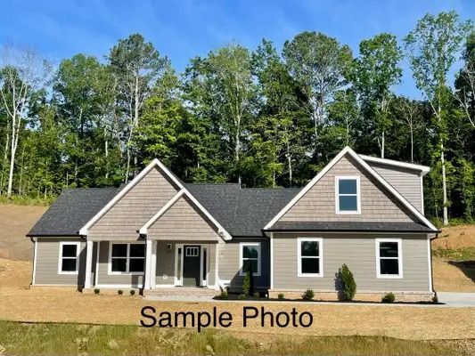 Single-story beige house with black roof and white trim, set in a wooded yard under a blue sky.