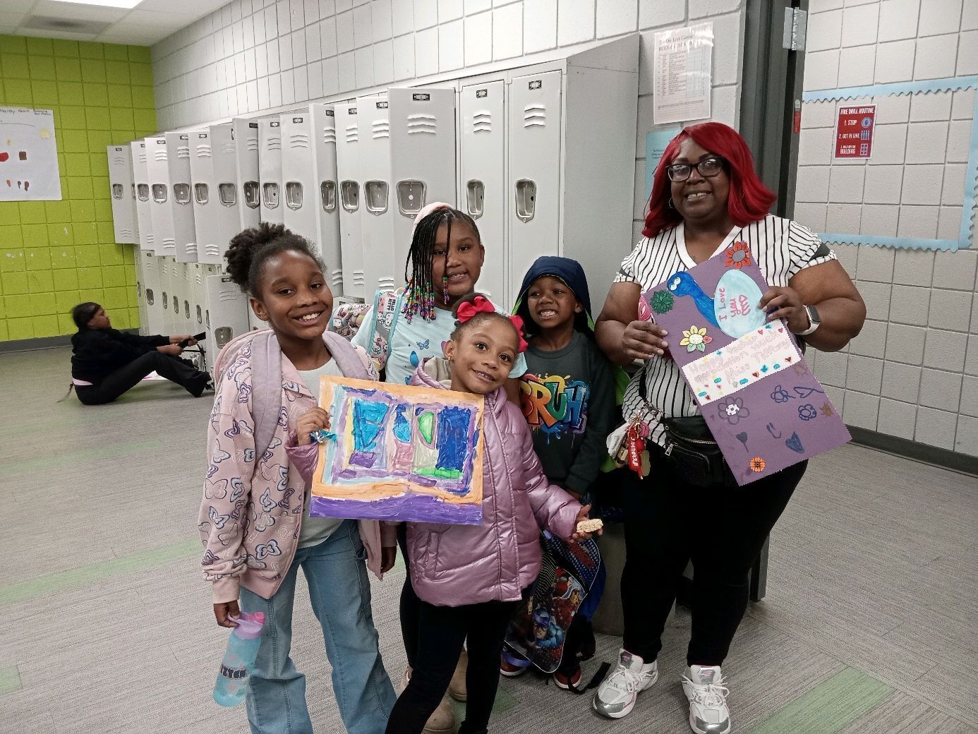 Students posing in a school hallway, holding colorful artwork and smiling in front of lockers.