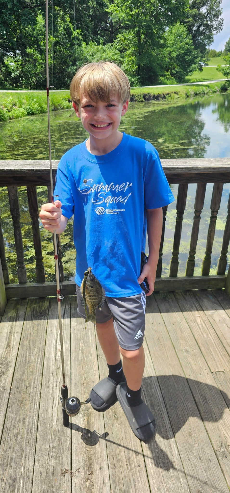 Smiling child in a blue shirt holding a fishing rod on a wooden deck by a pond