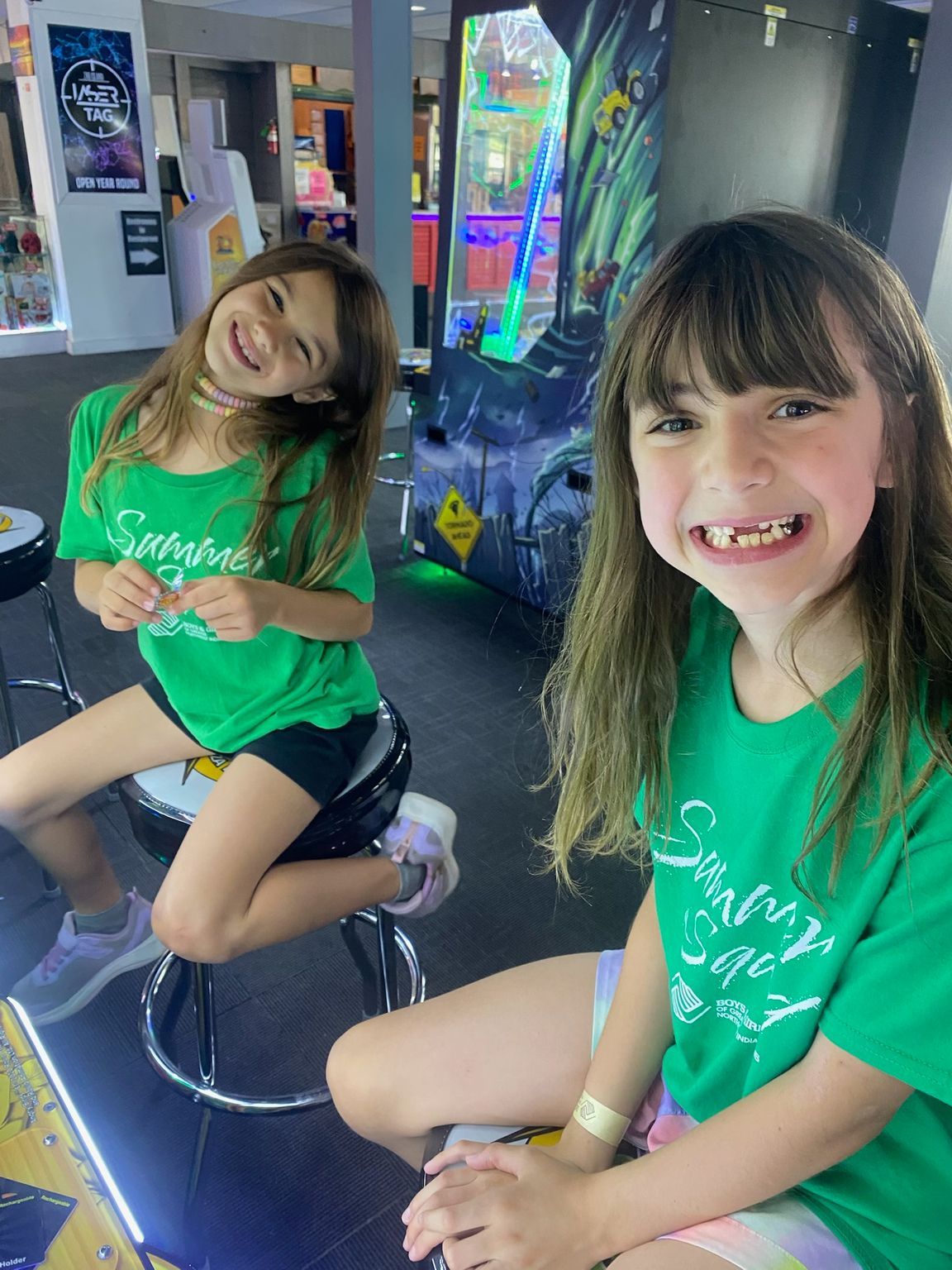 Two kids in green shirts smiling on stools in an arcade-like room