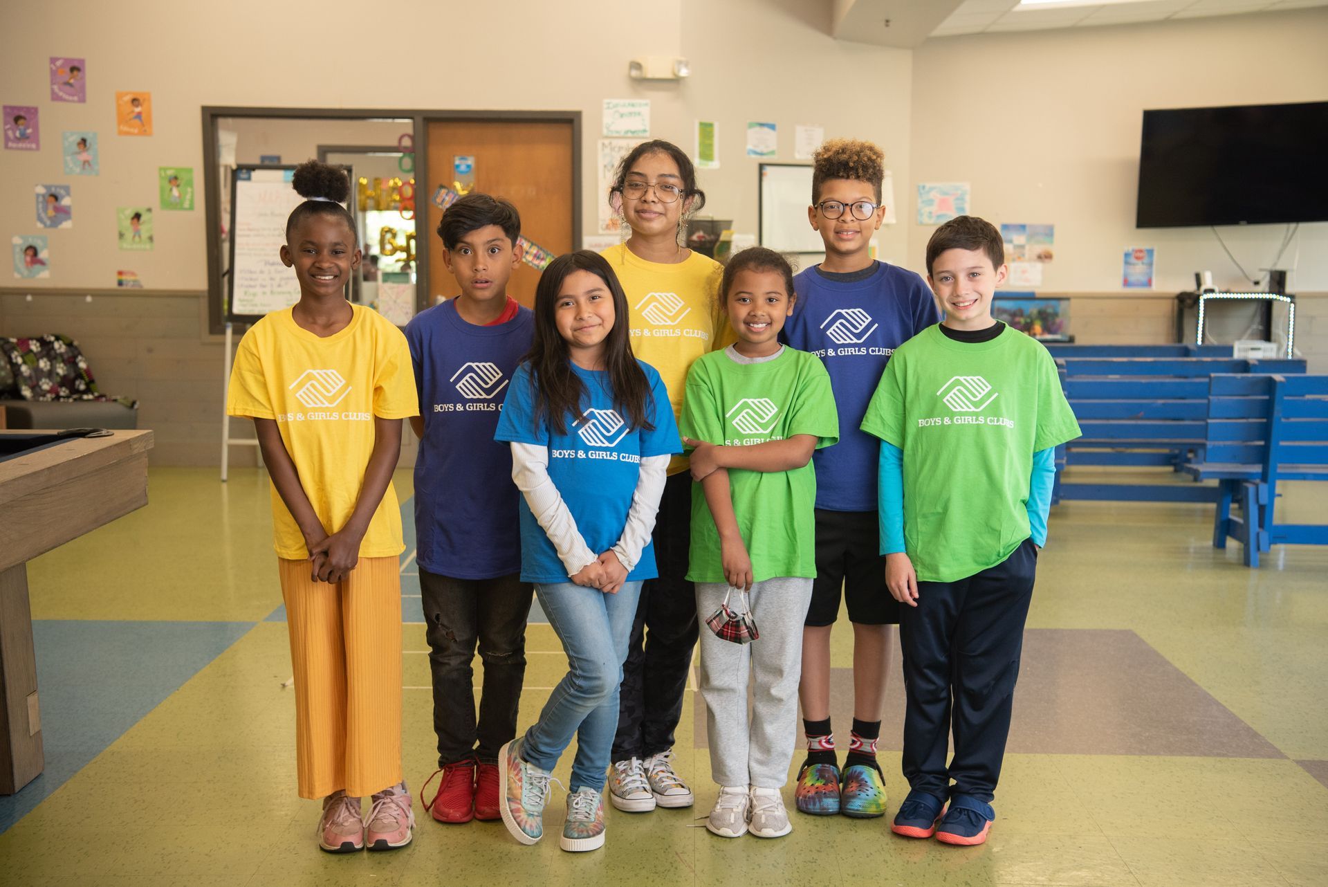 Seven children stand smiling in a classroom, wearing colorful matching T-shirts.