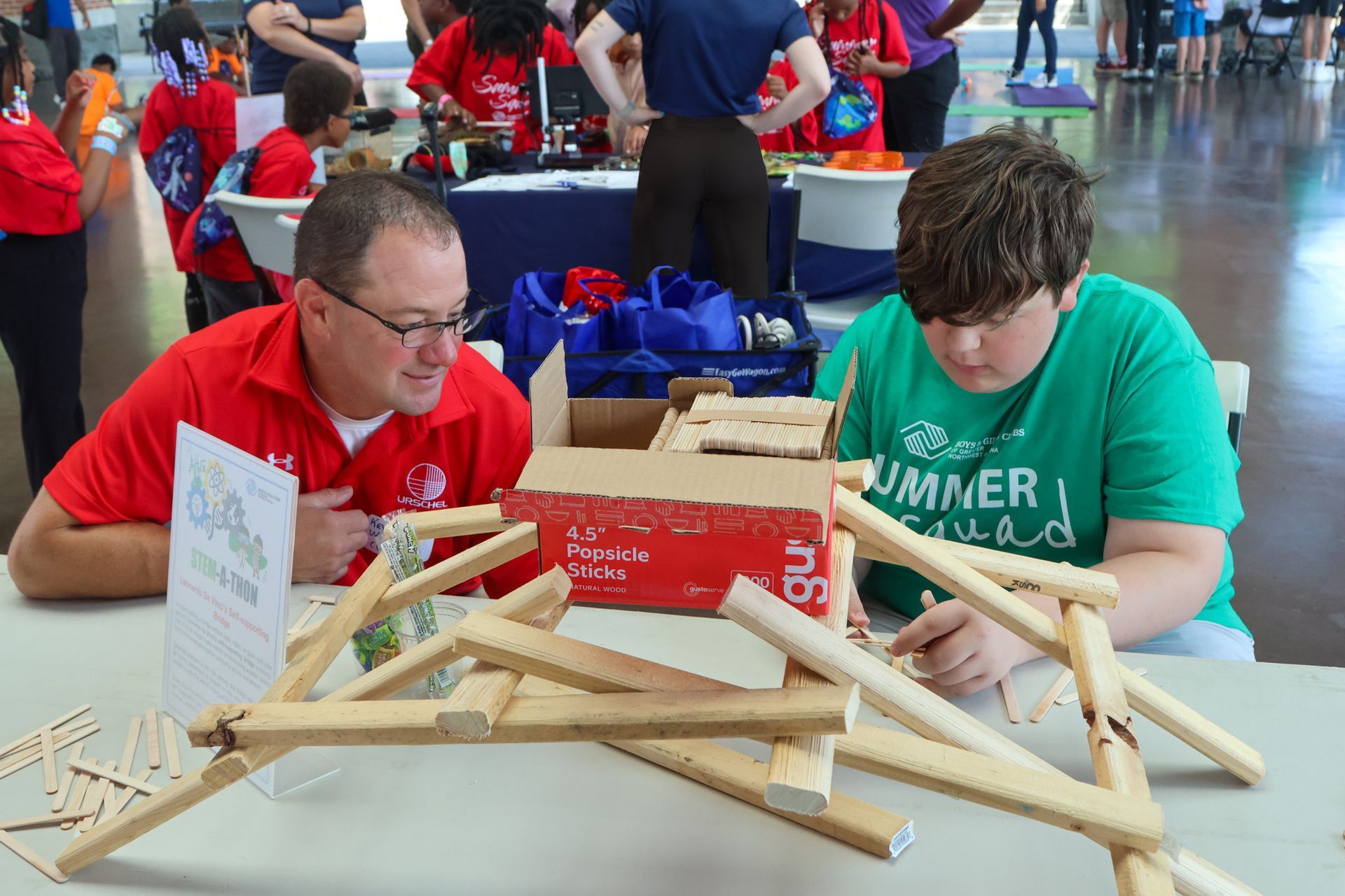 Two people lean over a table building a wooden structure at a crowded indoor event.