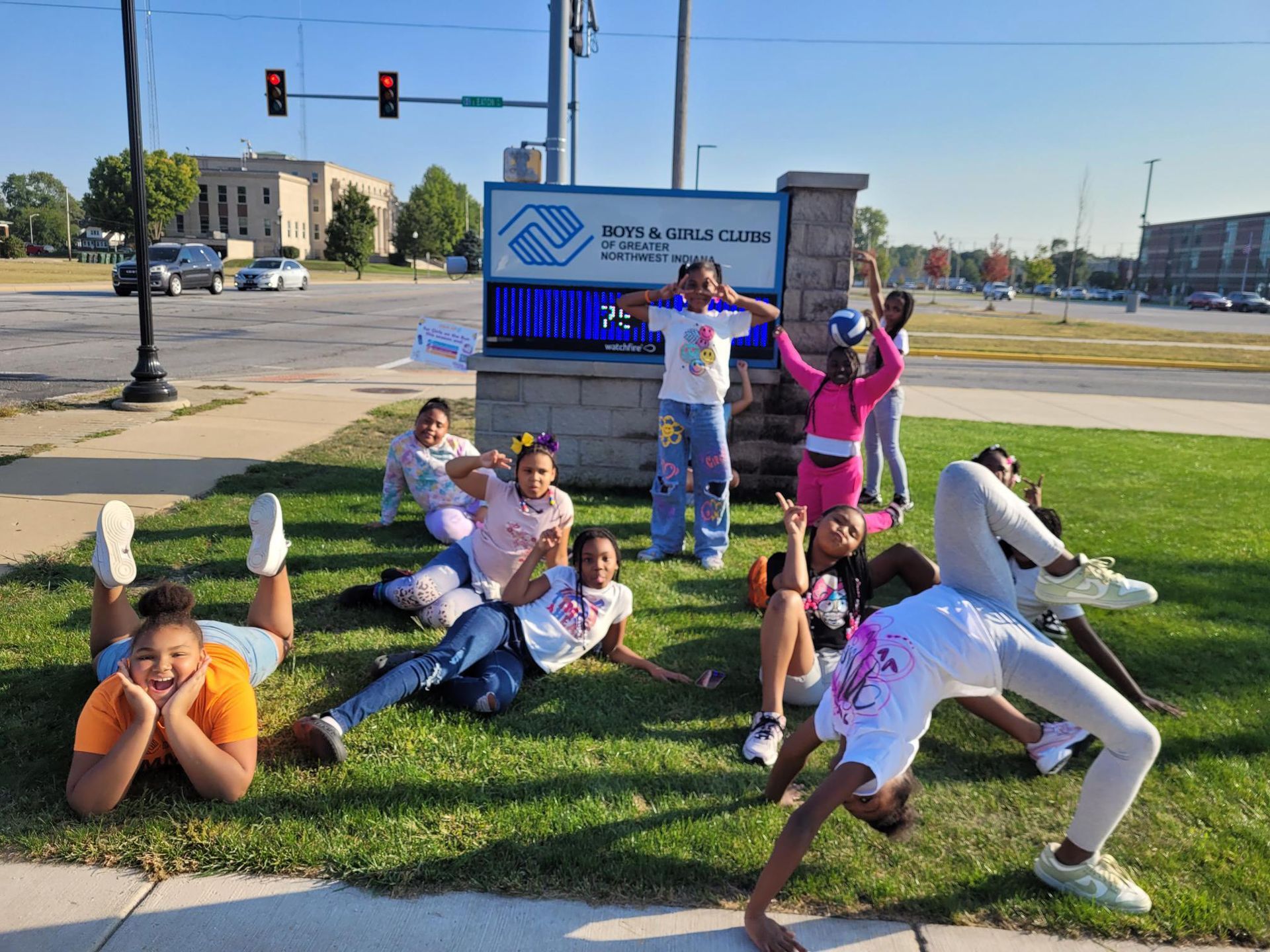 Group of people stretching on a grassy corner beside a road and blue sign on a sunny day