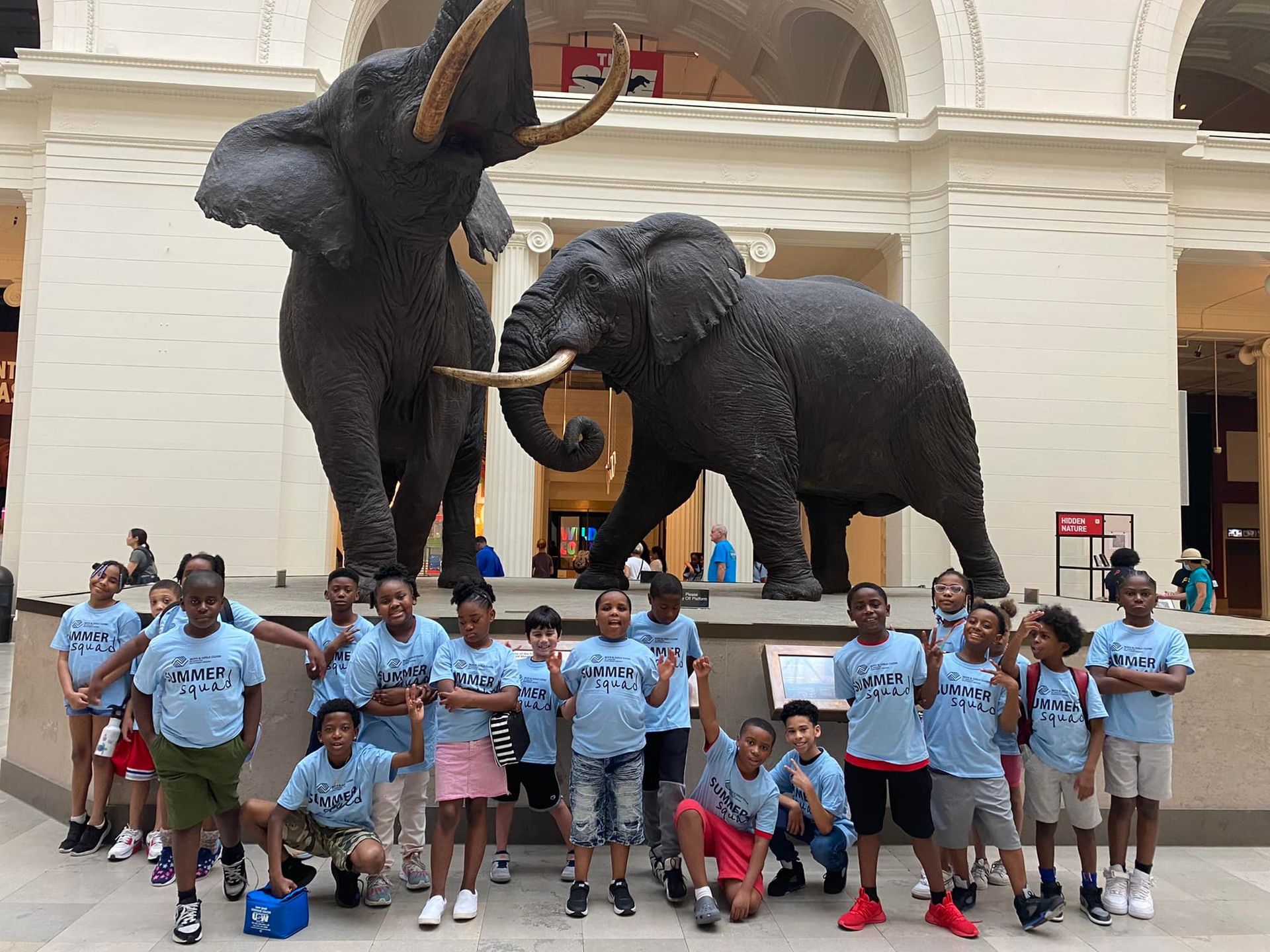 Group of children posing in blue shirts beneath two large elephant statues in a museum atrium