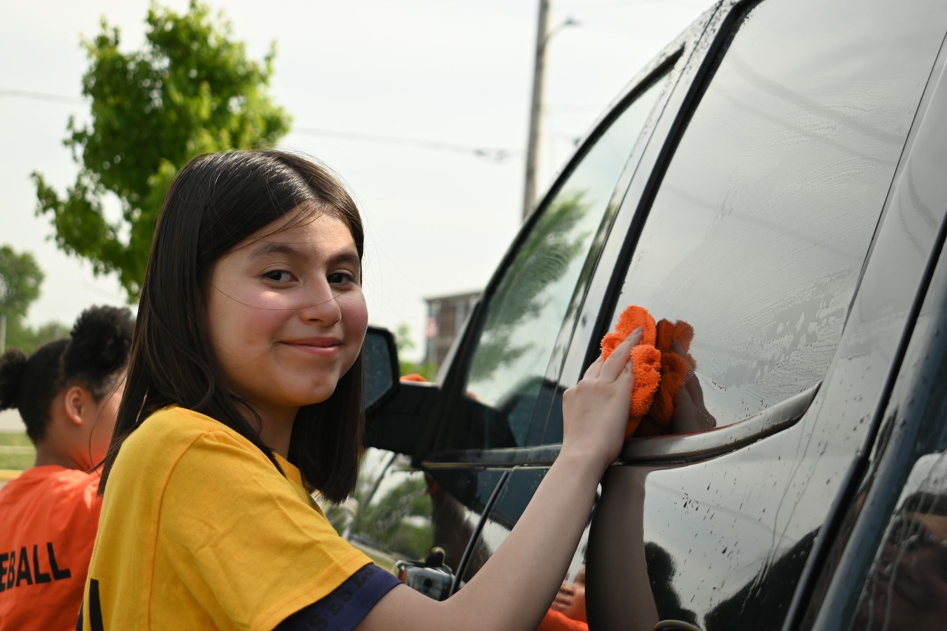 Person in a yellow shirt washing a black vehicle window with an orange cloth outdoors