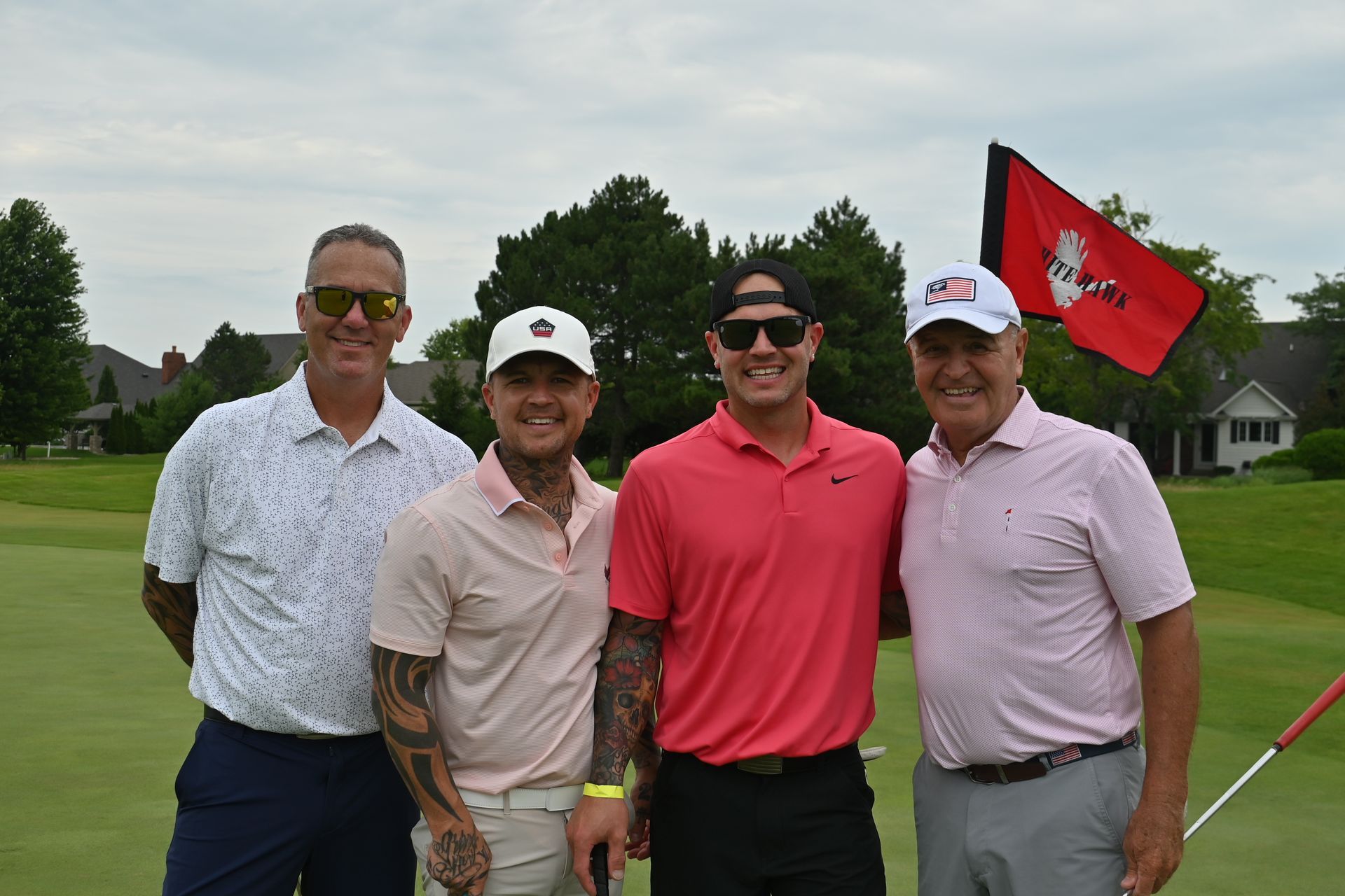Four men posing on a golf course, with a red flag and clubhouse in the background.