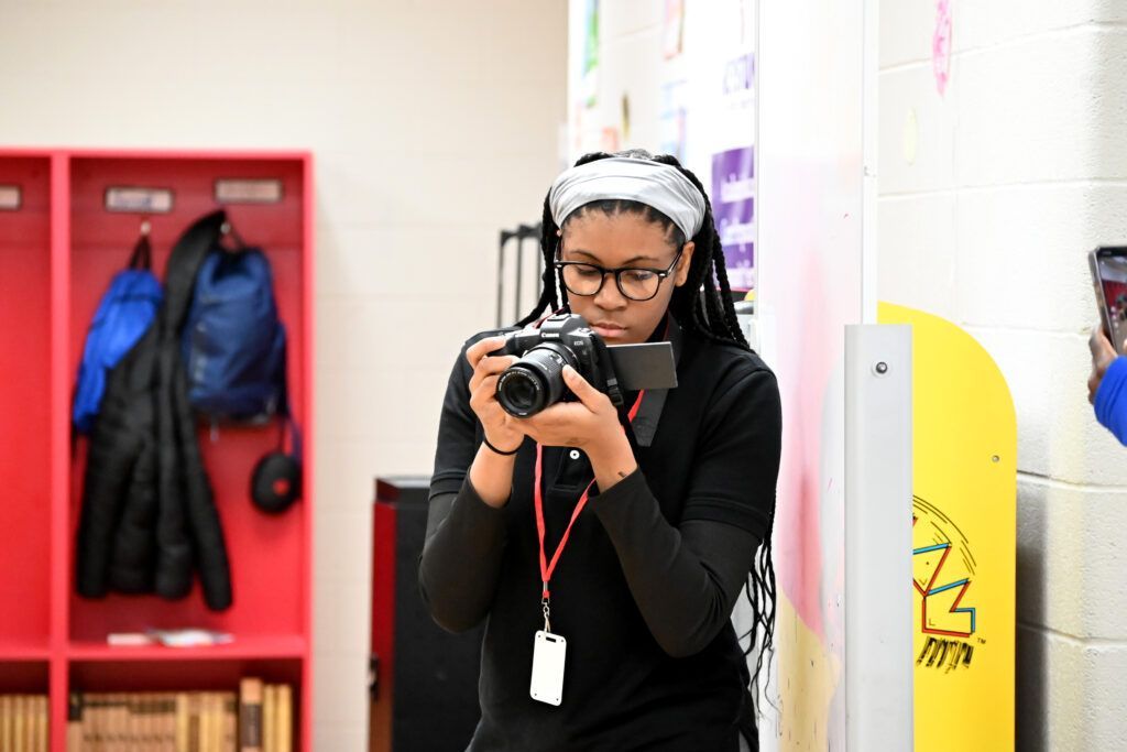 Person holding a camera in a classroom, with red lockers and a yellow bin in the background.