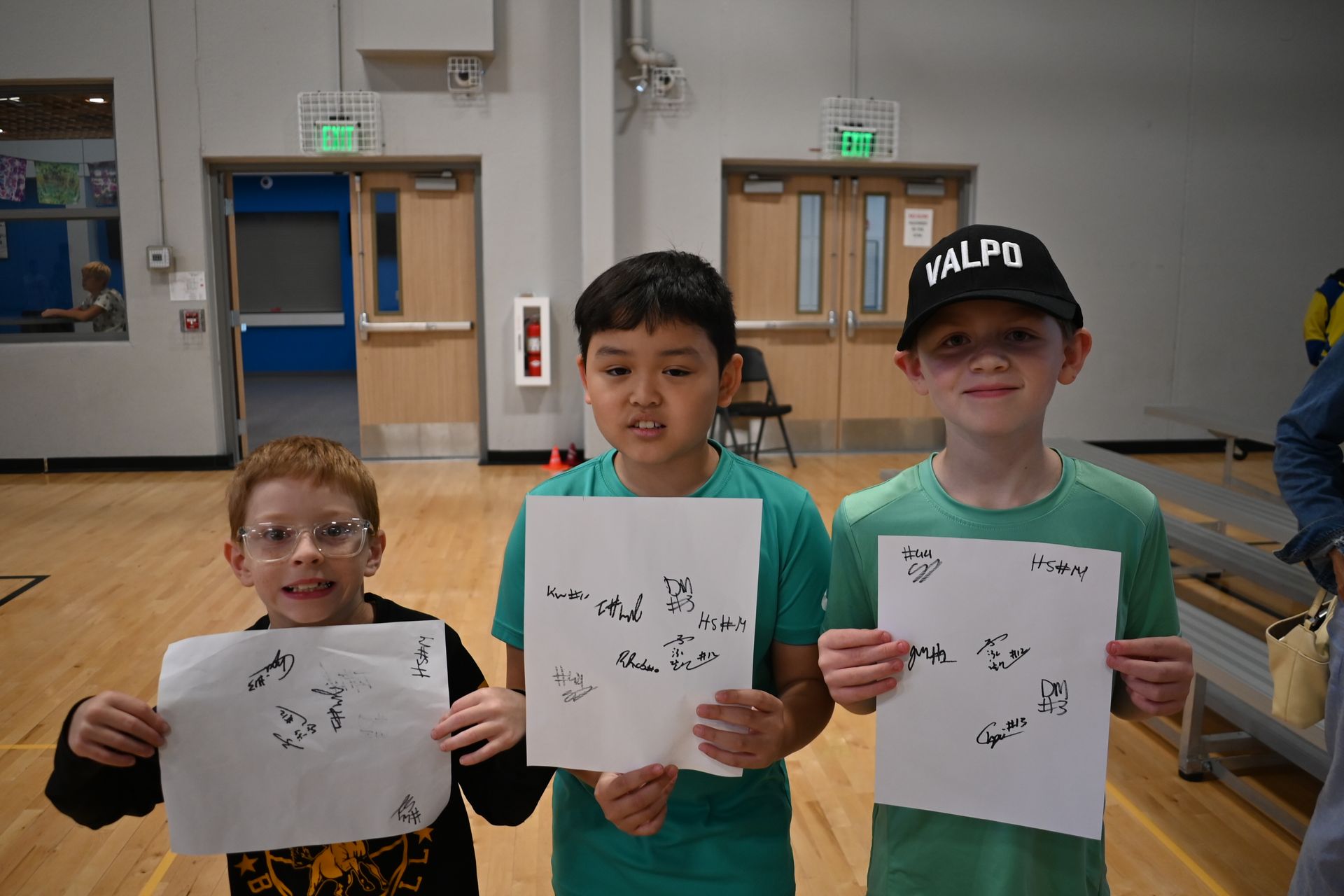 Three children in a gym hold up drawings on white paper.