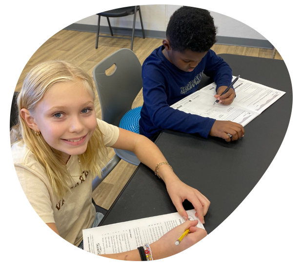 Two children working on worksheets at a table, one smiling at the camera.
