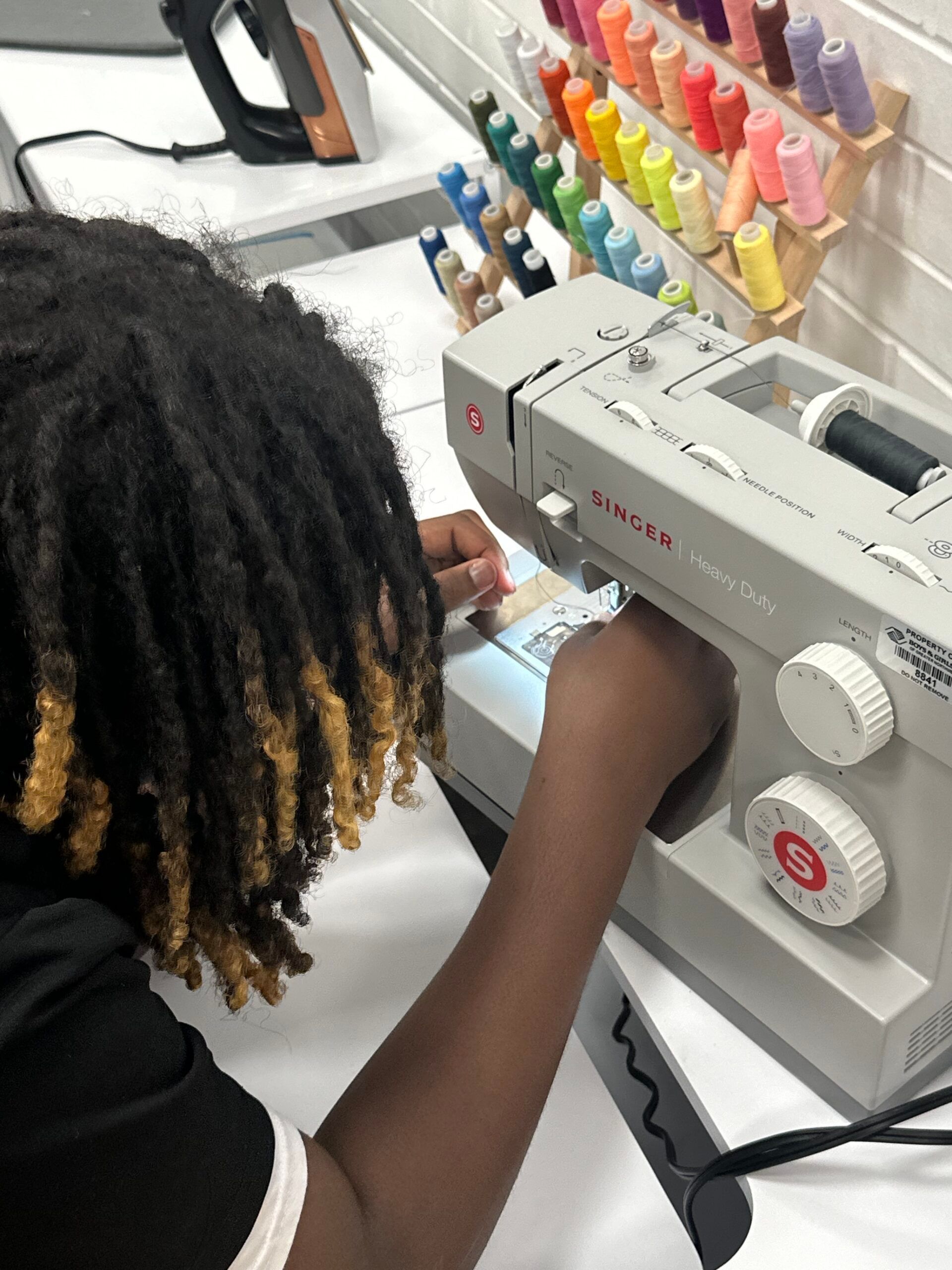 Person using a sewing machine beside a rack of colorful thread spools