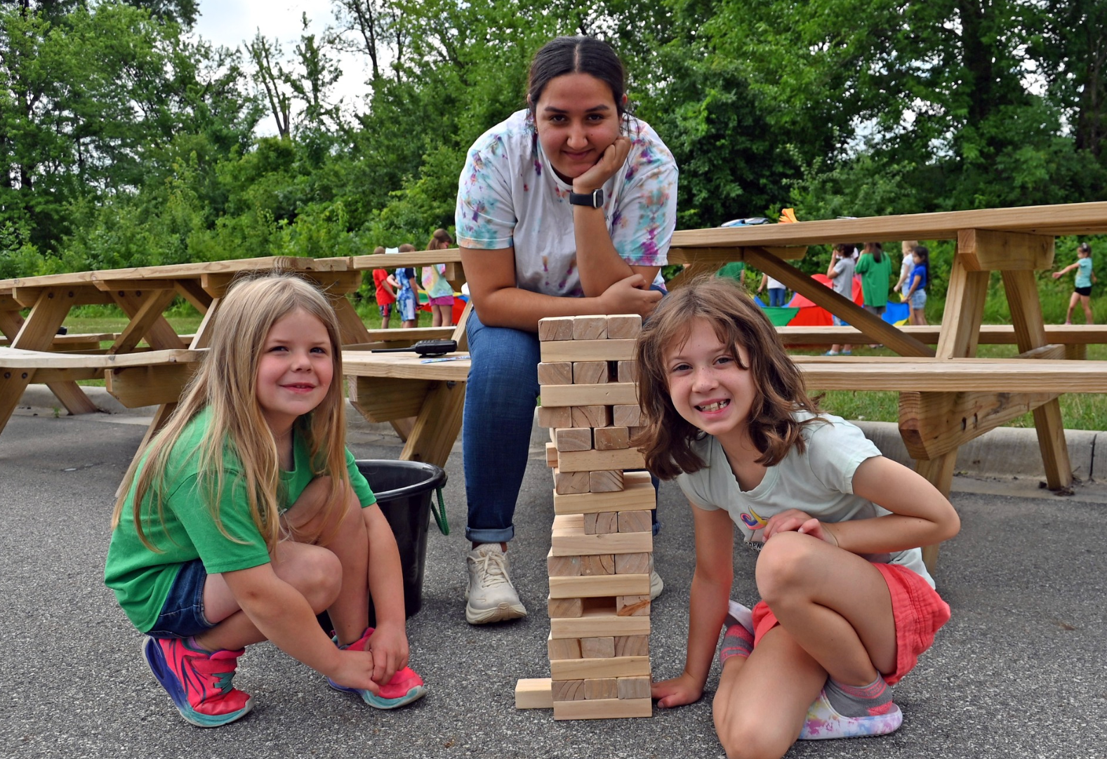 Three children crouch beside a tall wooden block tower outdoors on a picnic table area.