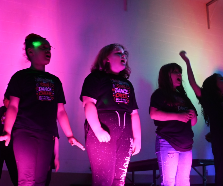 Four people on a pink-lit stage, posing with raised arms during a performance.