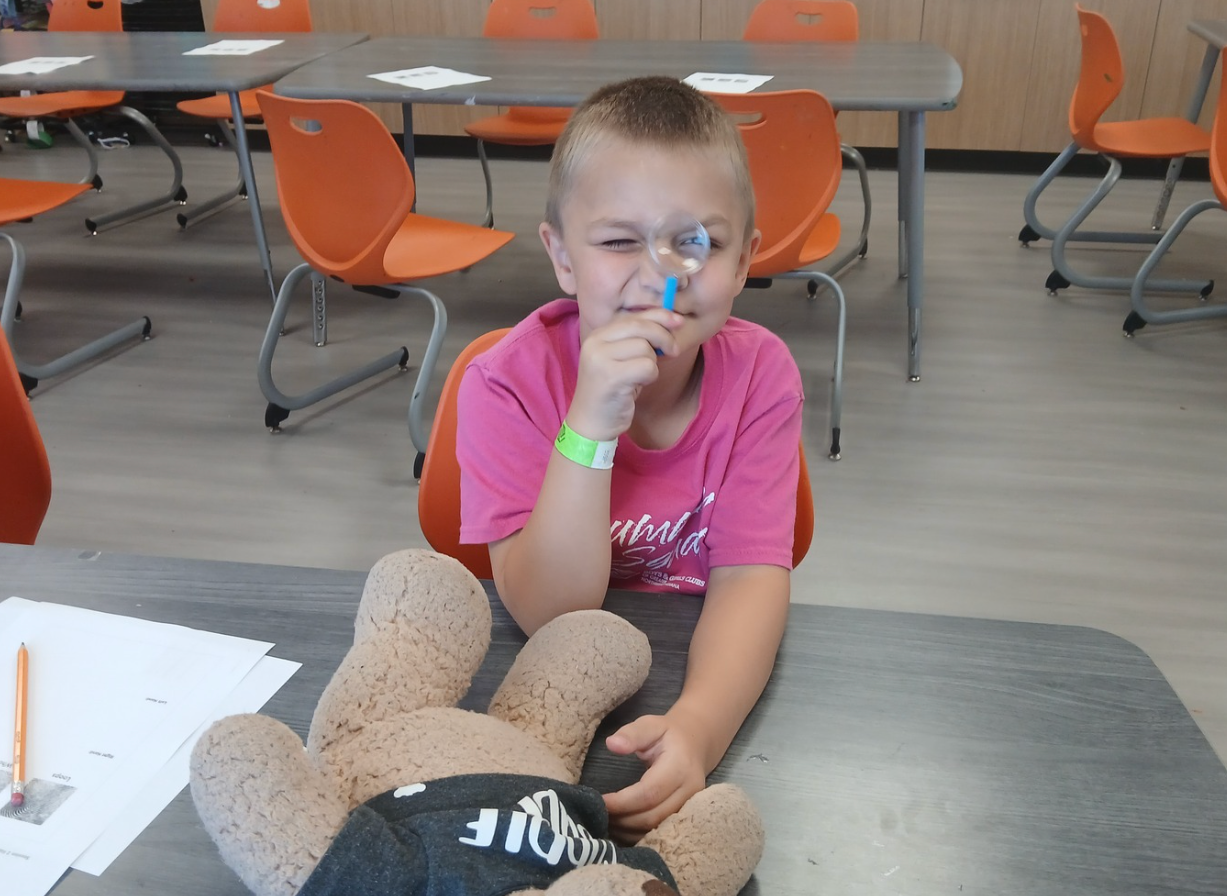 Child in a pink shirt smiling at a table in a classroom, with orange chairs in the background