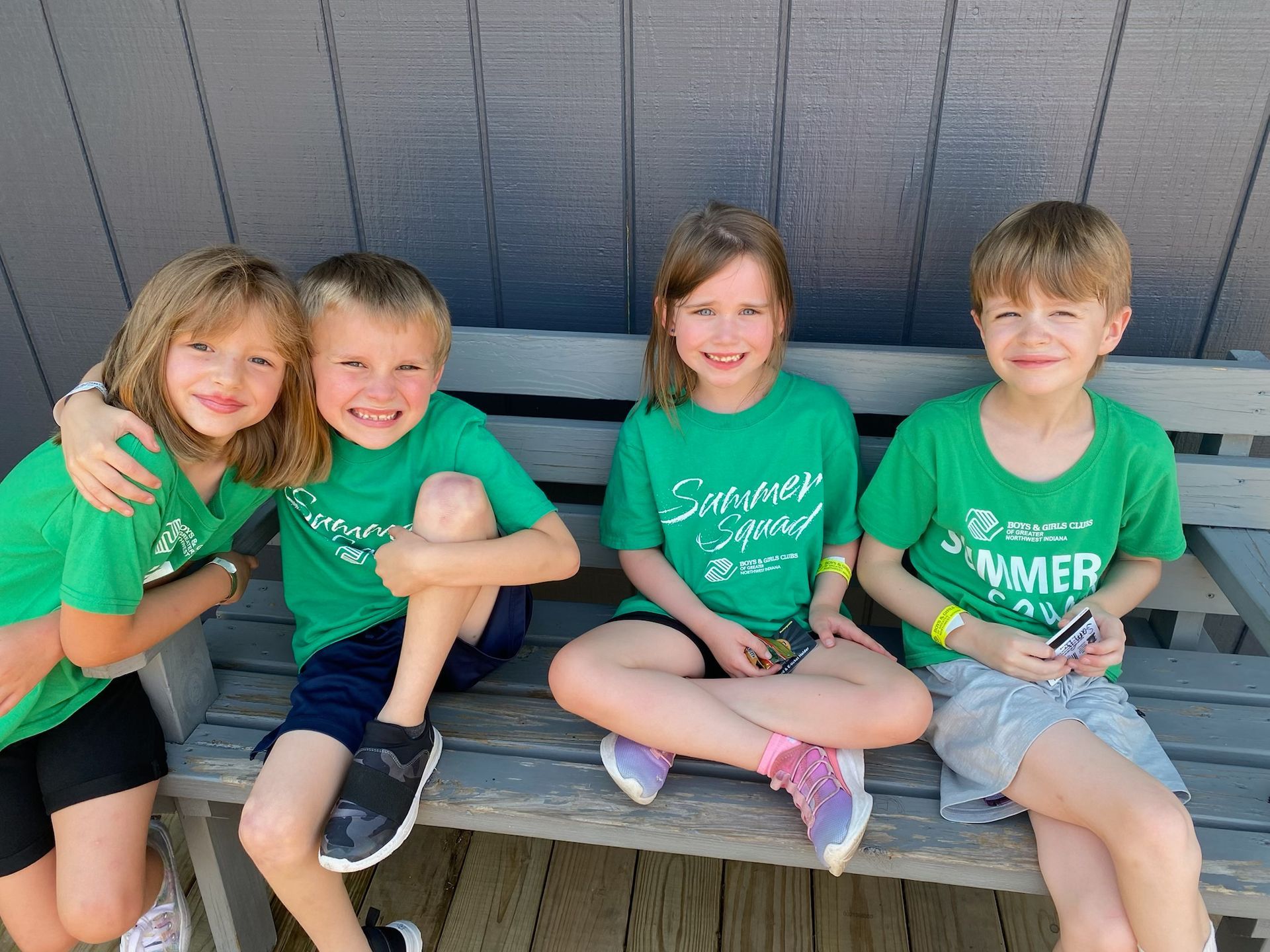 Four children in matching green shirts sit together on a bench, smiling at the camera.