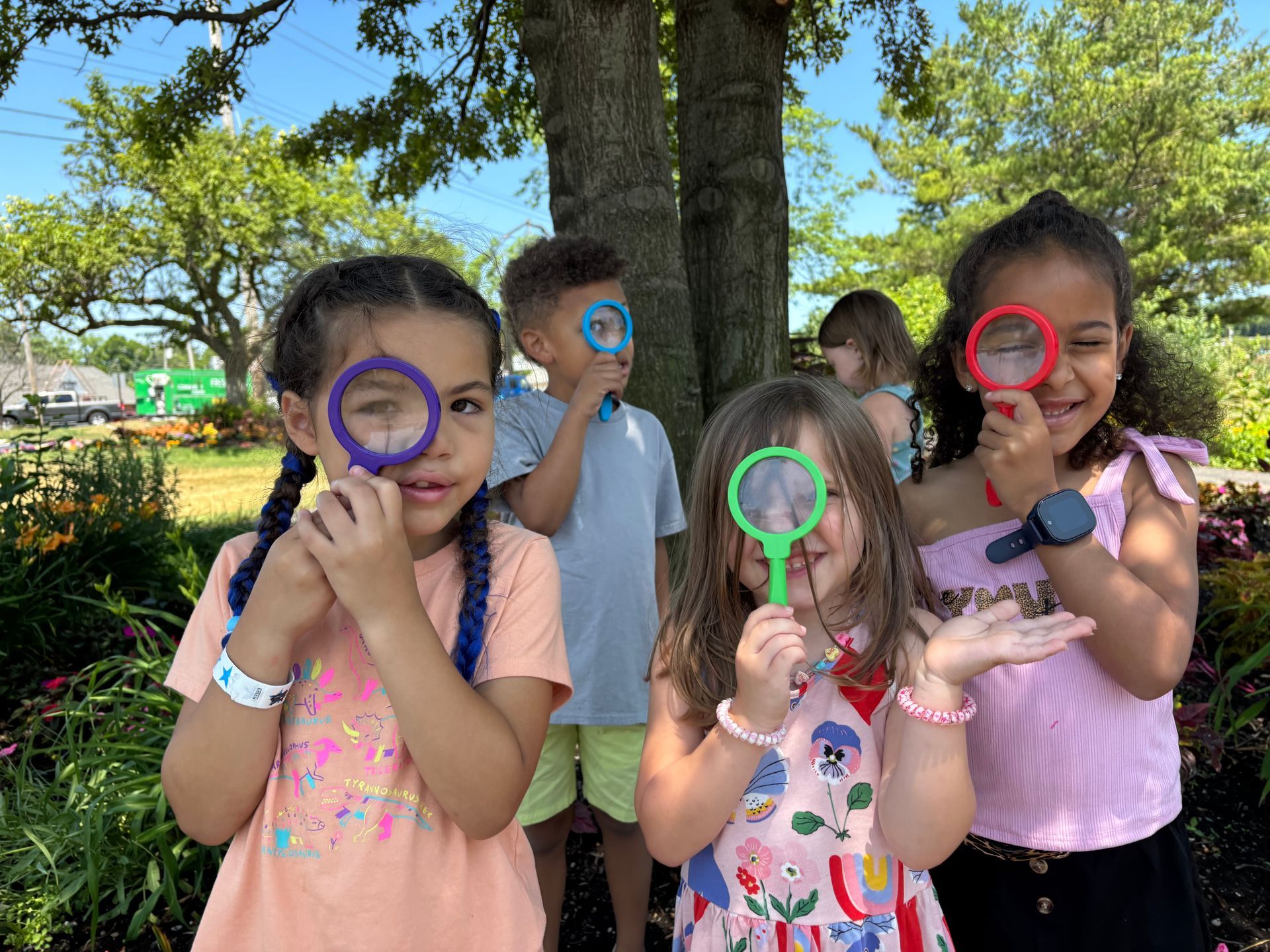 Four children outdoors holding colorful magnifying glasses under a tree, smiling at the camera