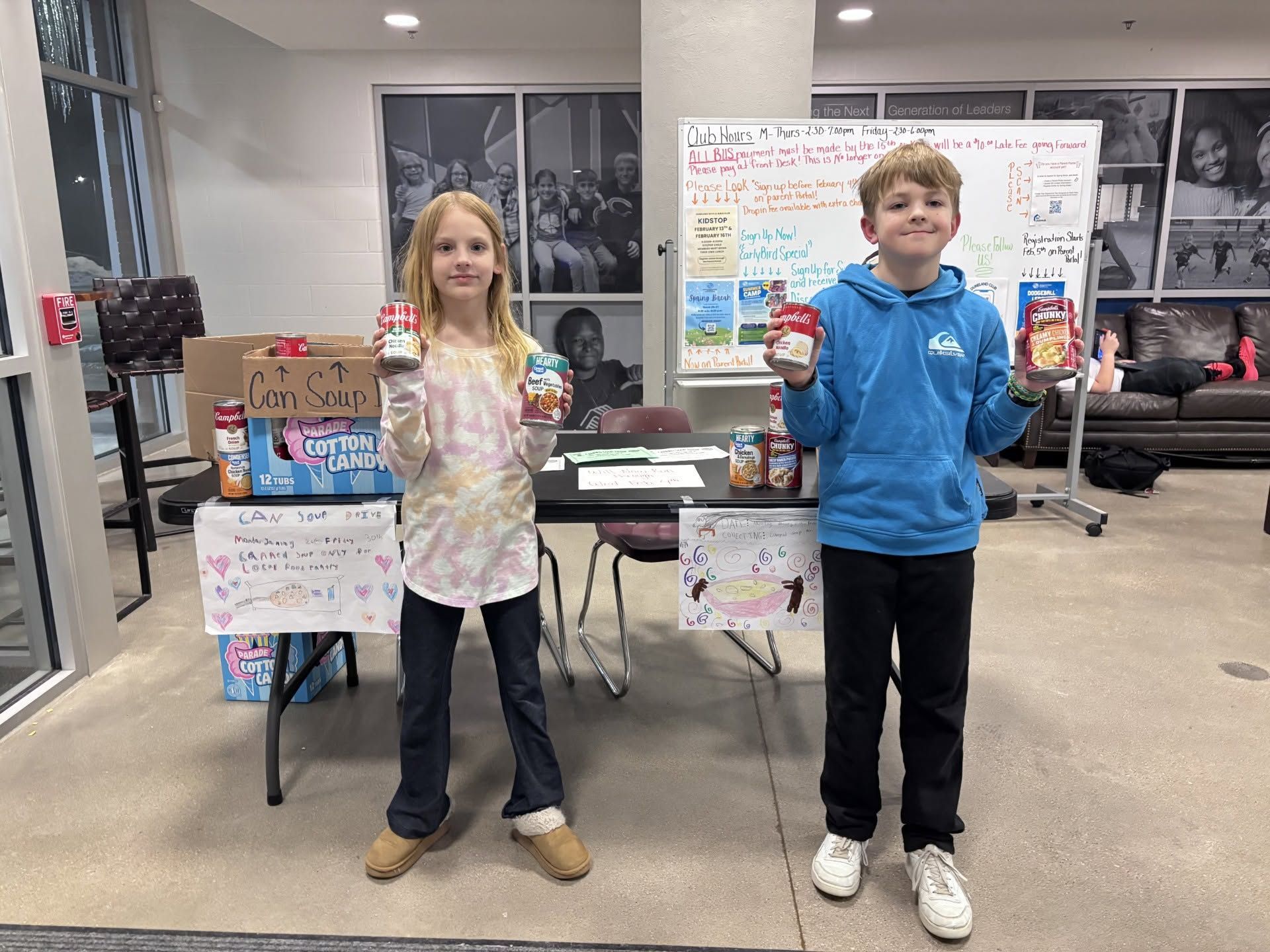 Two children in a classroom holding canned goods beside collection boxes and posters.