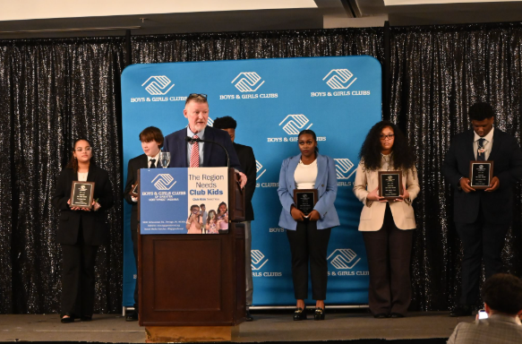Six people stand onstage at a podium during an awards presentation, with a blue backdrop behind them.