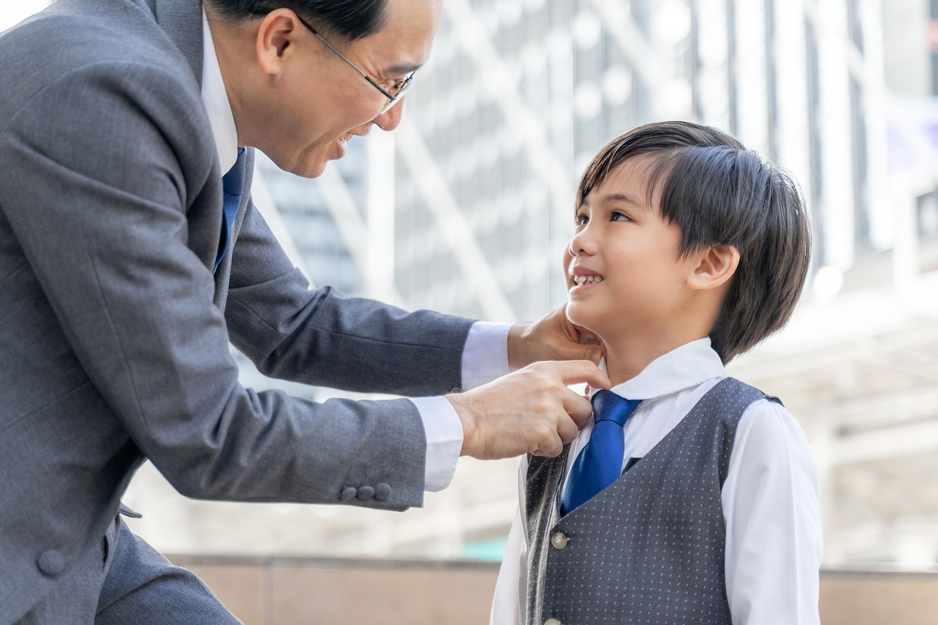 Man adjusting a boy’s tie outdoors, both smiling near a modern building