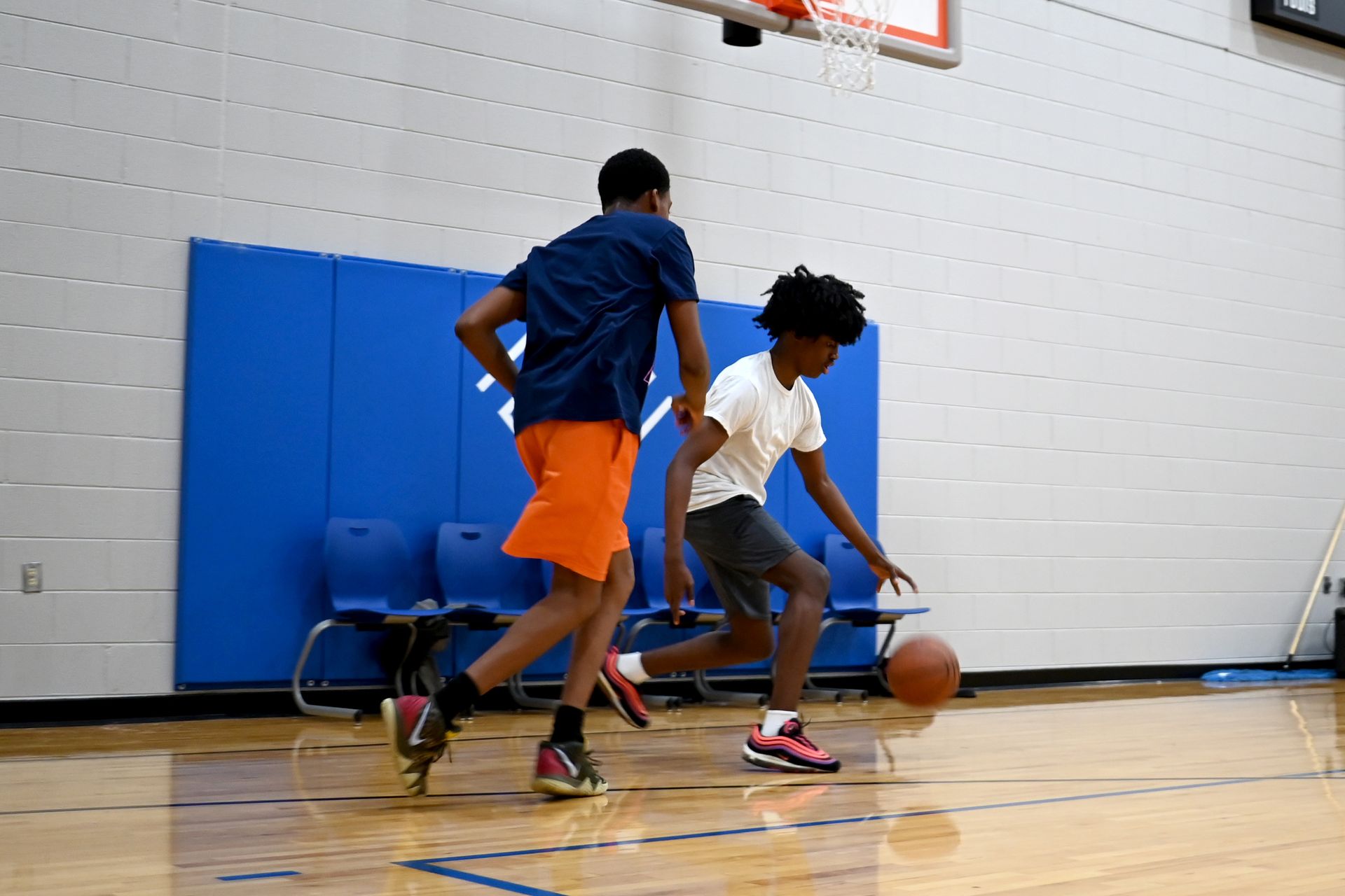 Two basketball players dribble on a gym court, with blue wall padding and a hoop in the background.