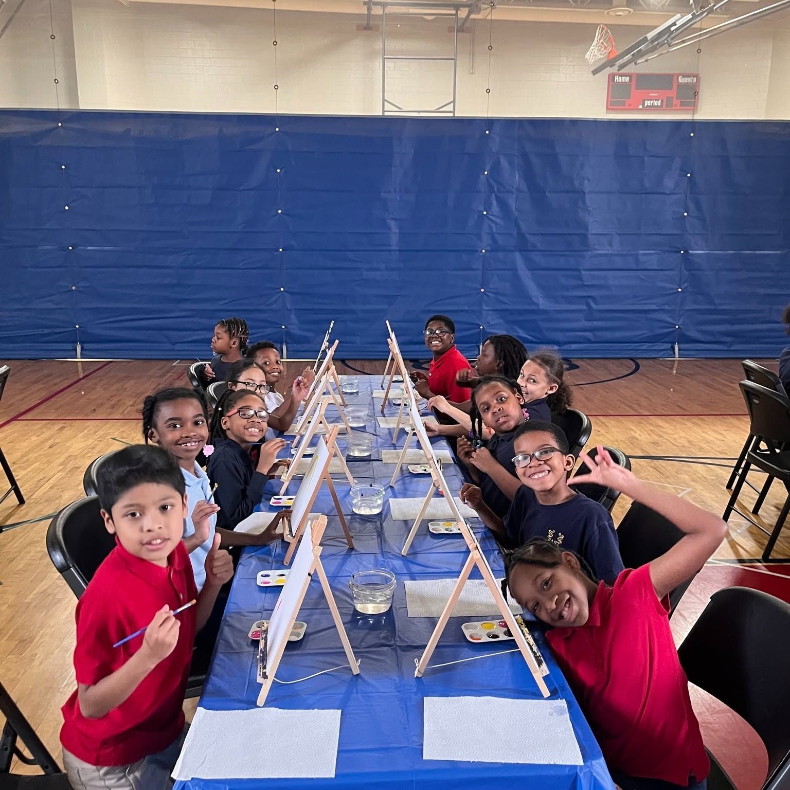 Students seated at a long table in a gym, smiling toward the camera with art supplies and small stands.