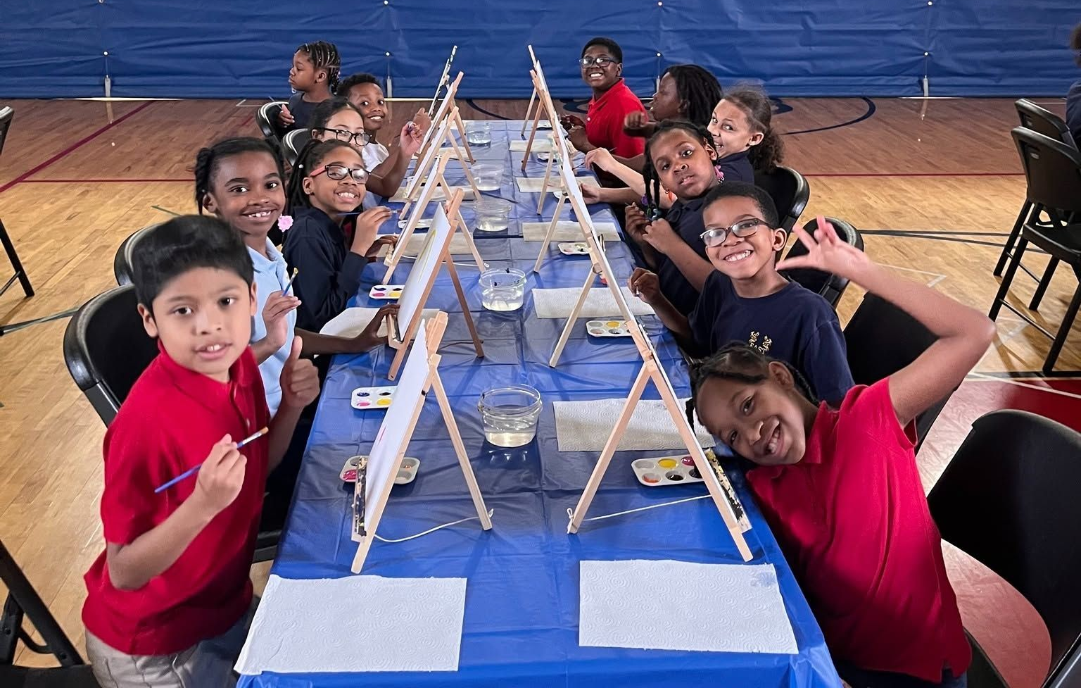 Children seated at a long blue banquet table, smiling in a gymnasium with a central display setup