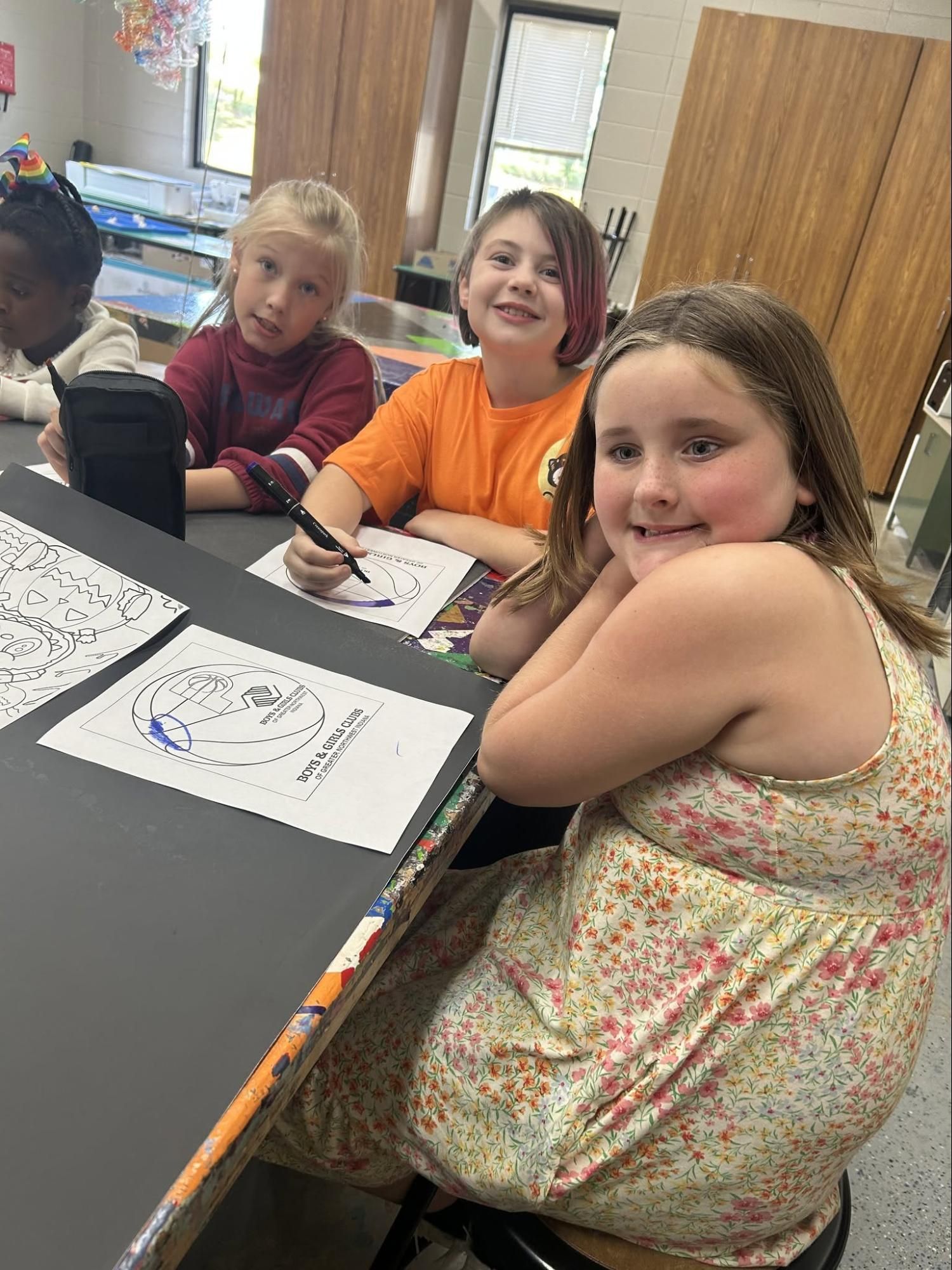 Children seated at a classroom table, smiling and working on papers with markers