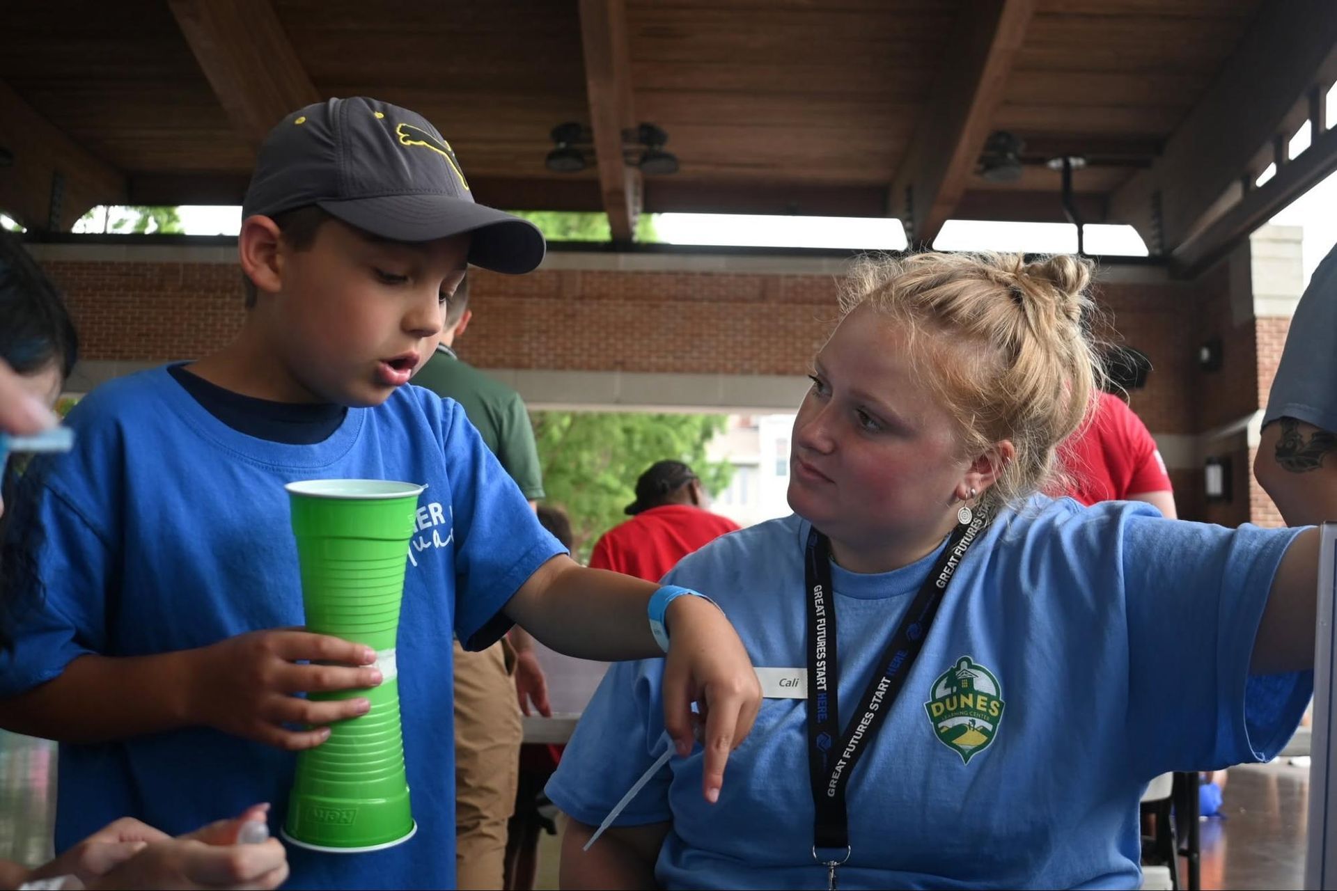 Two children in blue shirts talk at a picnic table, one holding a bright green cup.