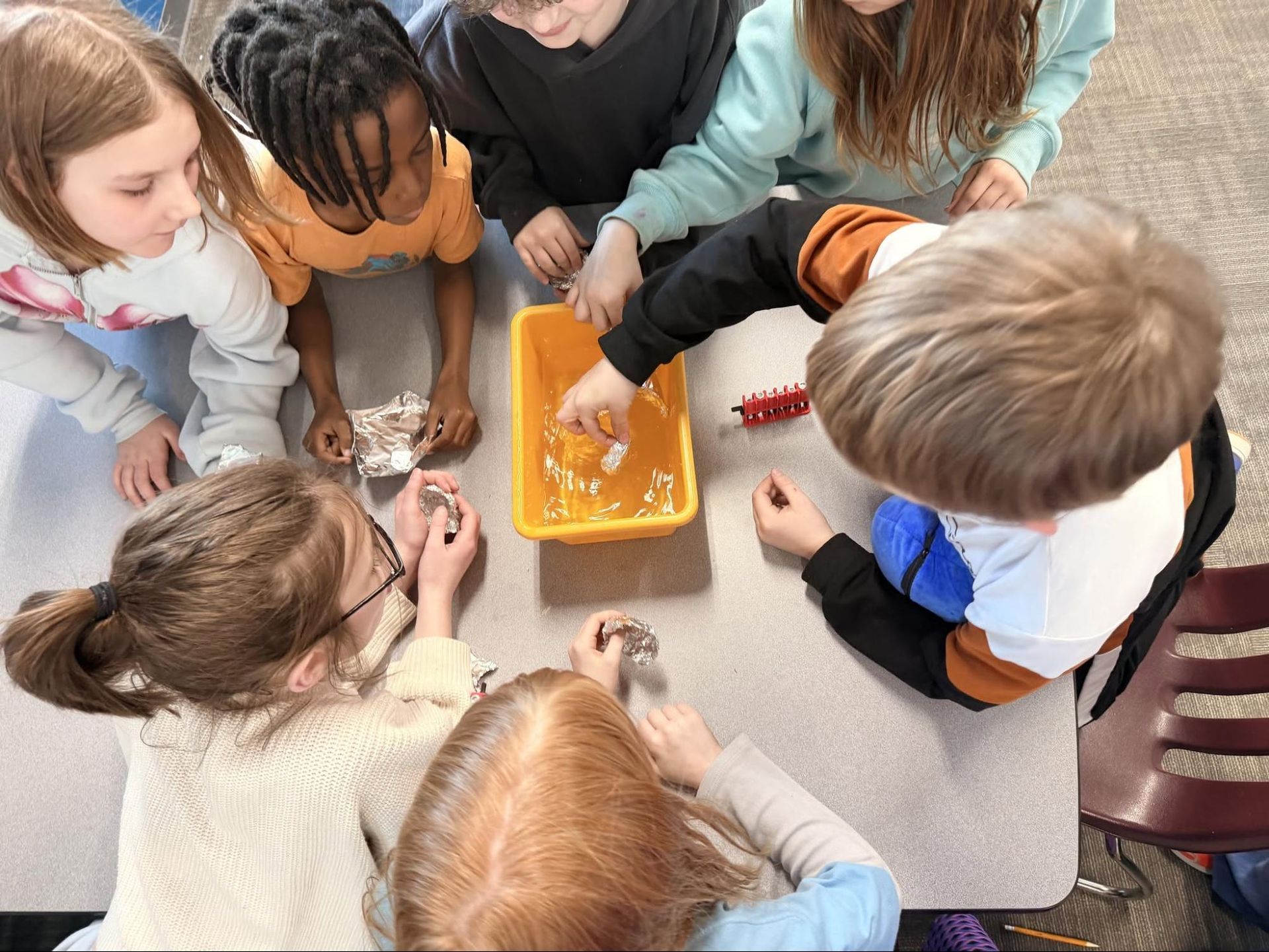 Children gather around a table, watching hands sort small items in a yellow tray.