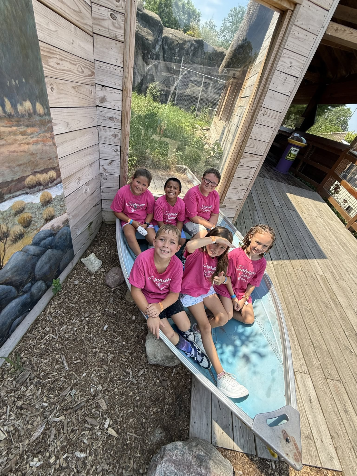 Five people in pink shirts sit on a small platform beside a wooden building and stone wall.
