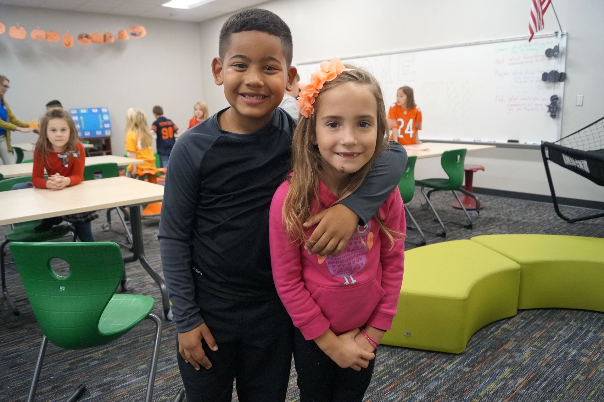 Two children smiling in a classroom, with colorful seating and other students in the background