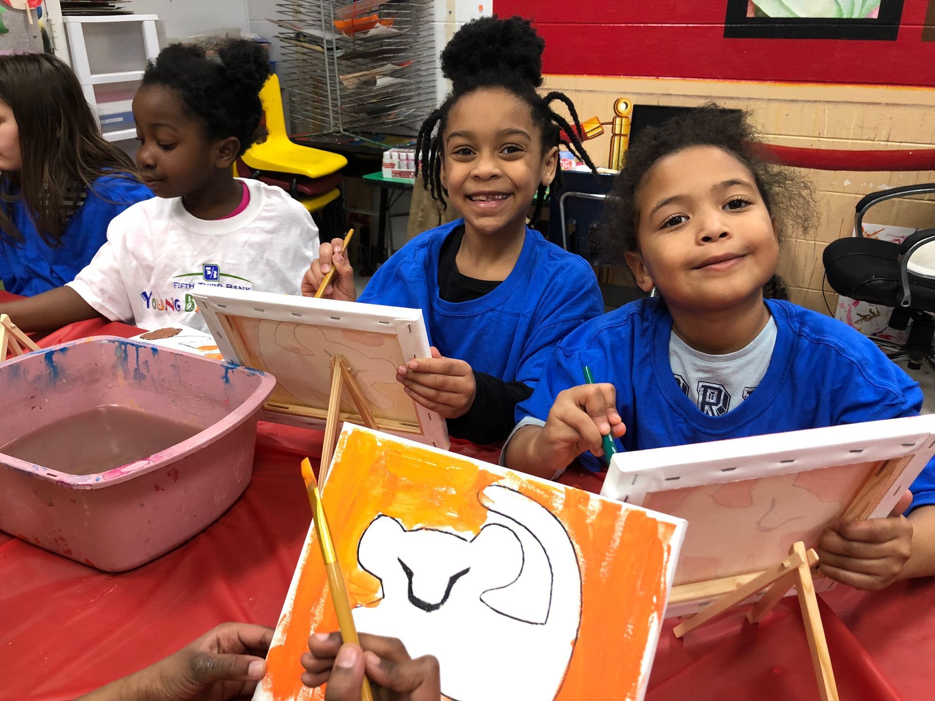 Children painting at a classroom table with brushes and canvases, smiling in front of an orange art project.