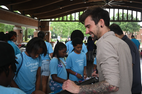 Man in beige shirt smiling and handing items to a group of people in blue shirts under a pavilion