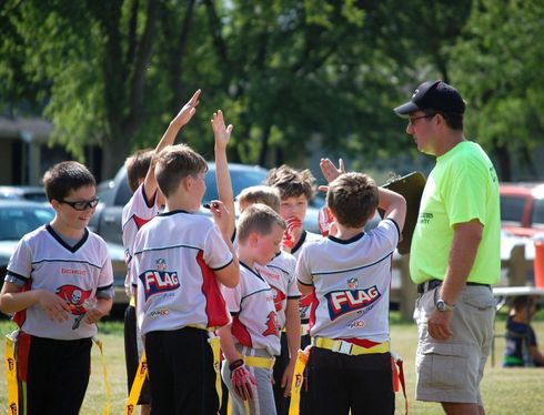 Youth flag football team huddles with coach on a sunny field, raising hands before a game.