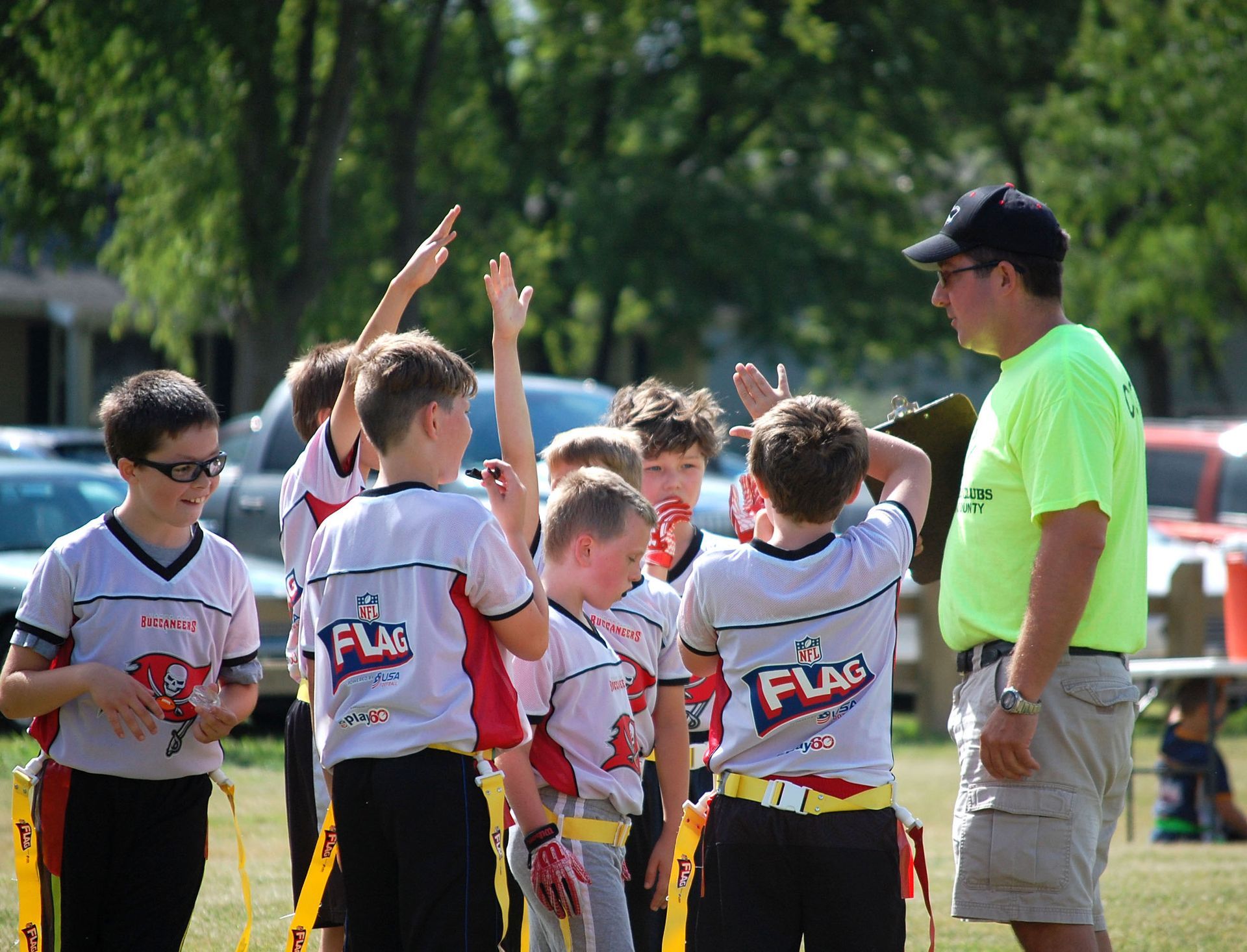 Youth flag football team huddles with coach on a sunny field, players raising hands in white and red jerseys