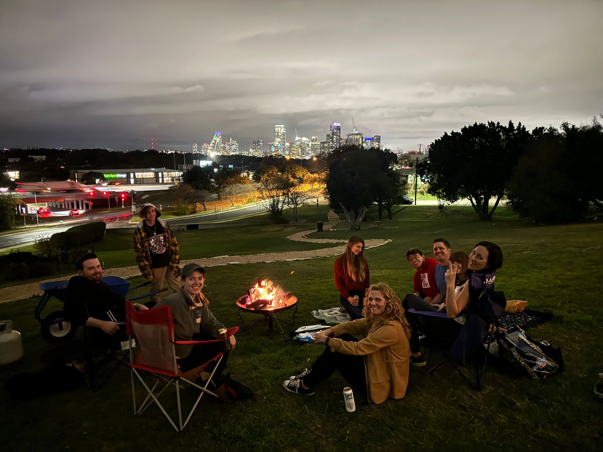 Friends gathered around a campfire on a grassy hill at night with city lights in the distance
