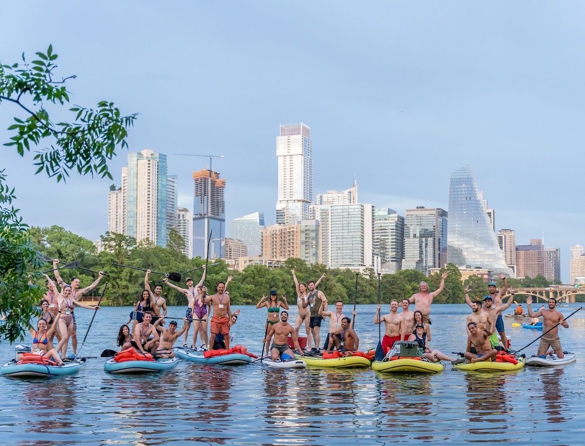 People on colorful paddleboards on a river with a city skyline in the background