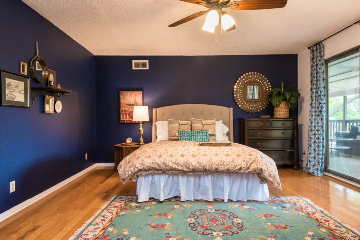 Spacious bedroom with navy walls, a queen bed, patterned rug, ceiling fan, and dresser by the window.