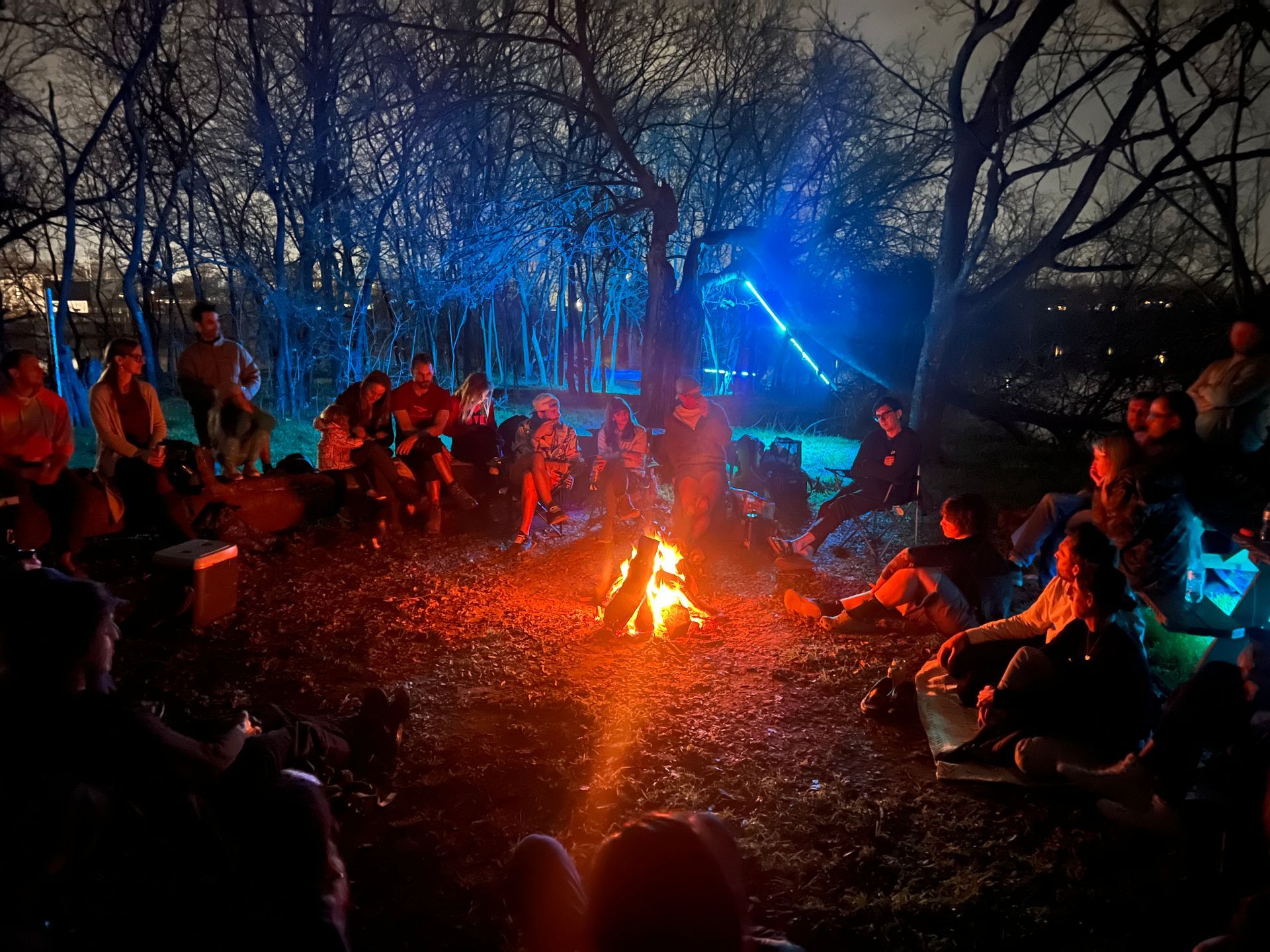 People gathered around a campfire in a dark wooded area, with blue light glowing in the background.