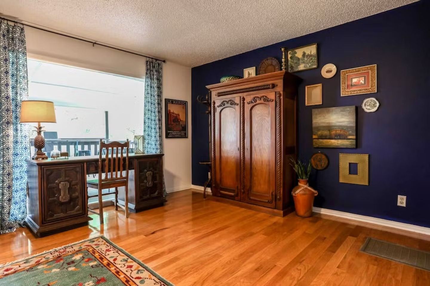 Living room with wood floors, navy accent wall, large armoire, desk by window, and patterned rug