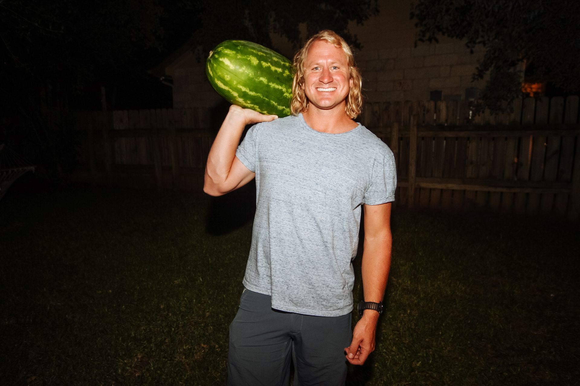 Person in a gray shirt holding a large watermelon on their shoulder outdoors at night.