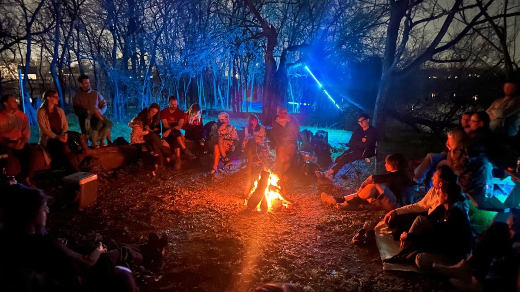 People seated around a campfire in a dark wooded area, lit by blue stage lights.