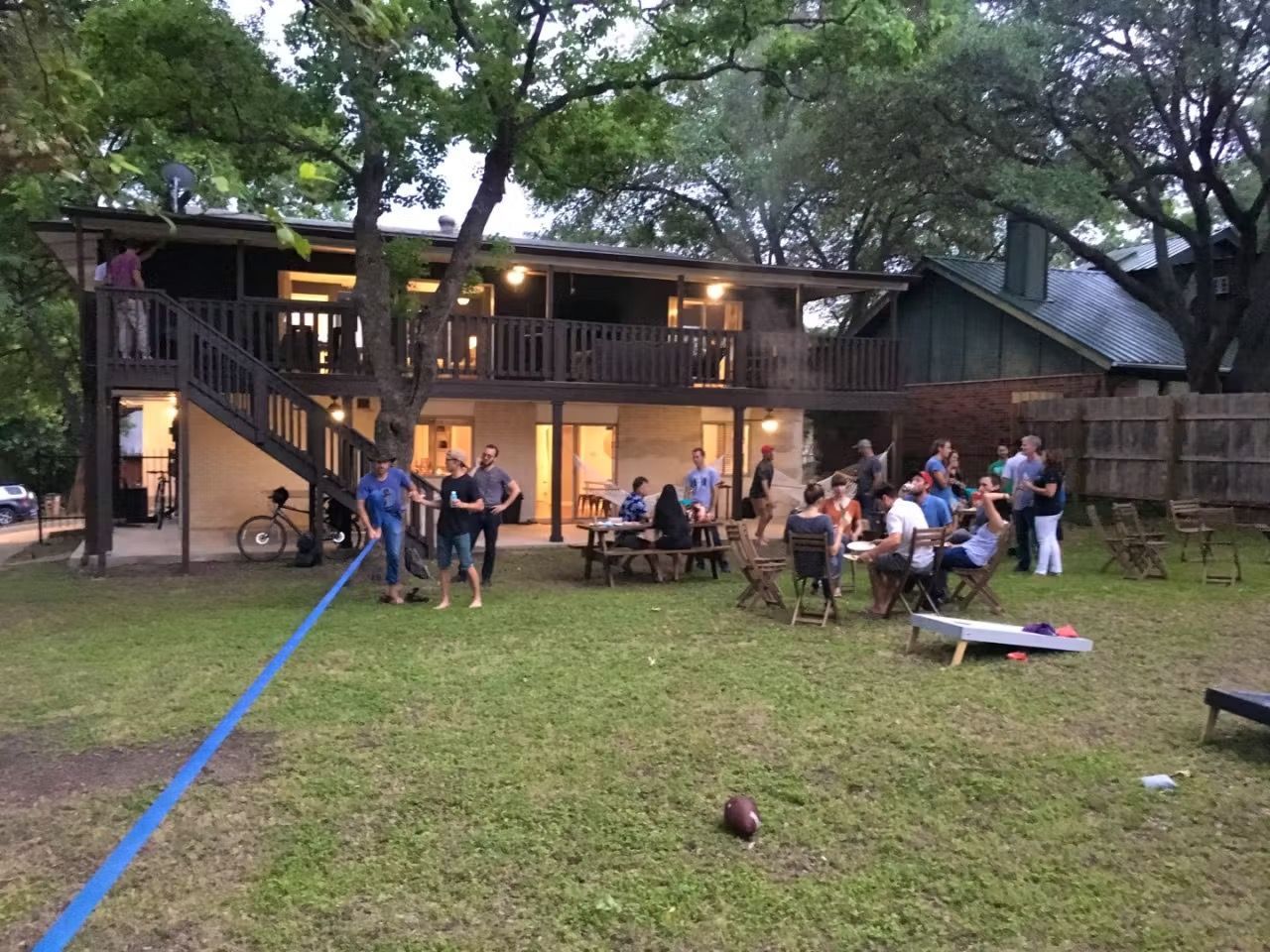 People gathered outside a wooden lodge at dusk, sitting and standing on a grassy lawn.