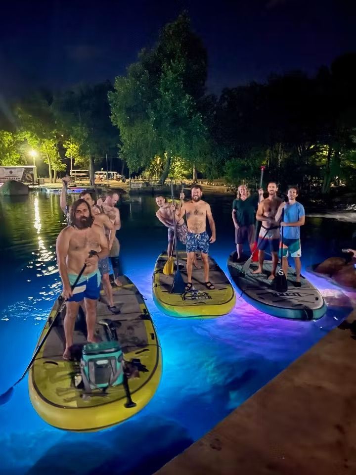 People standing on paddleboards at night under blue lights beside a calm waterfront
