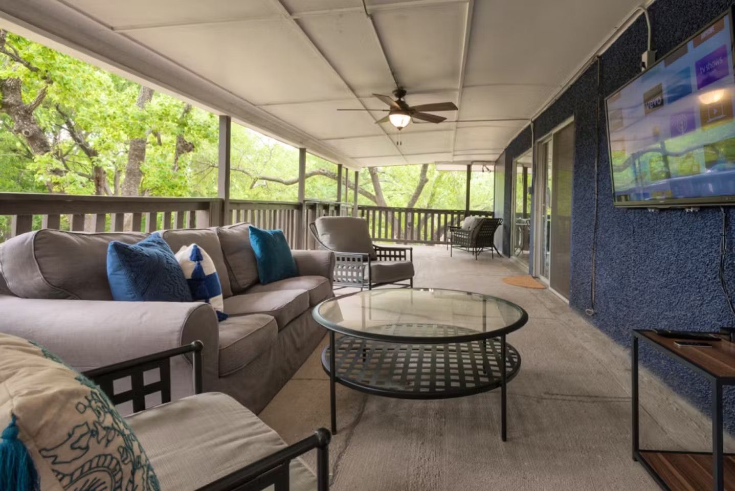Covered patio with sectional sofa, glass coffee table, TV, and ceiling fan overlooking greenery