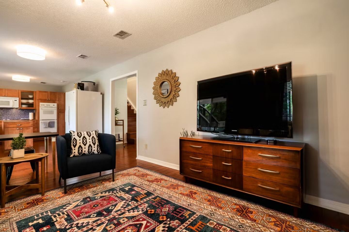 Living room with patterned rug, black chair, large TV, and wooden dresser near kitchen doorway