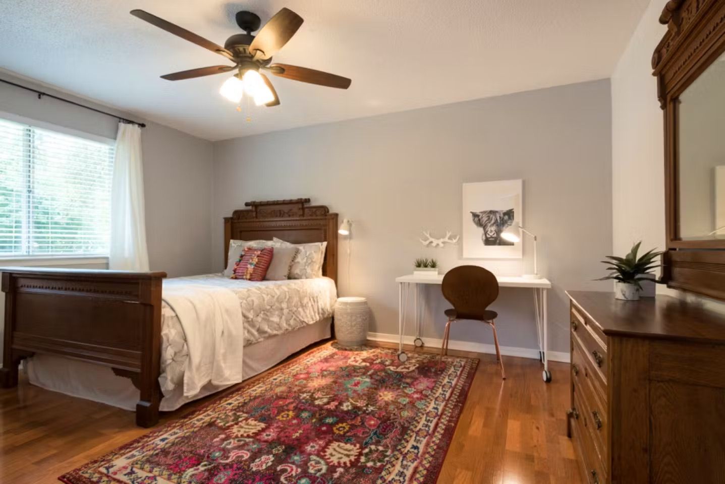Bright bedroom with wooden bed, patterned rug, desk, and ceiling fan under a window.