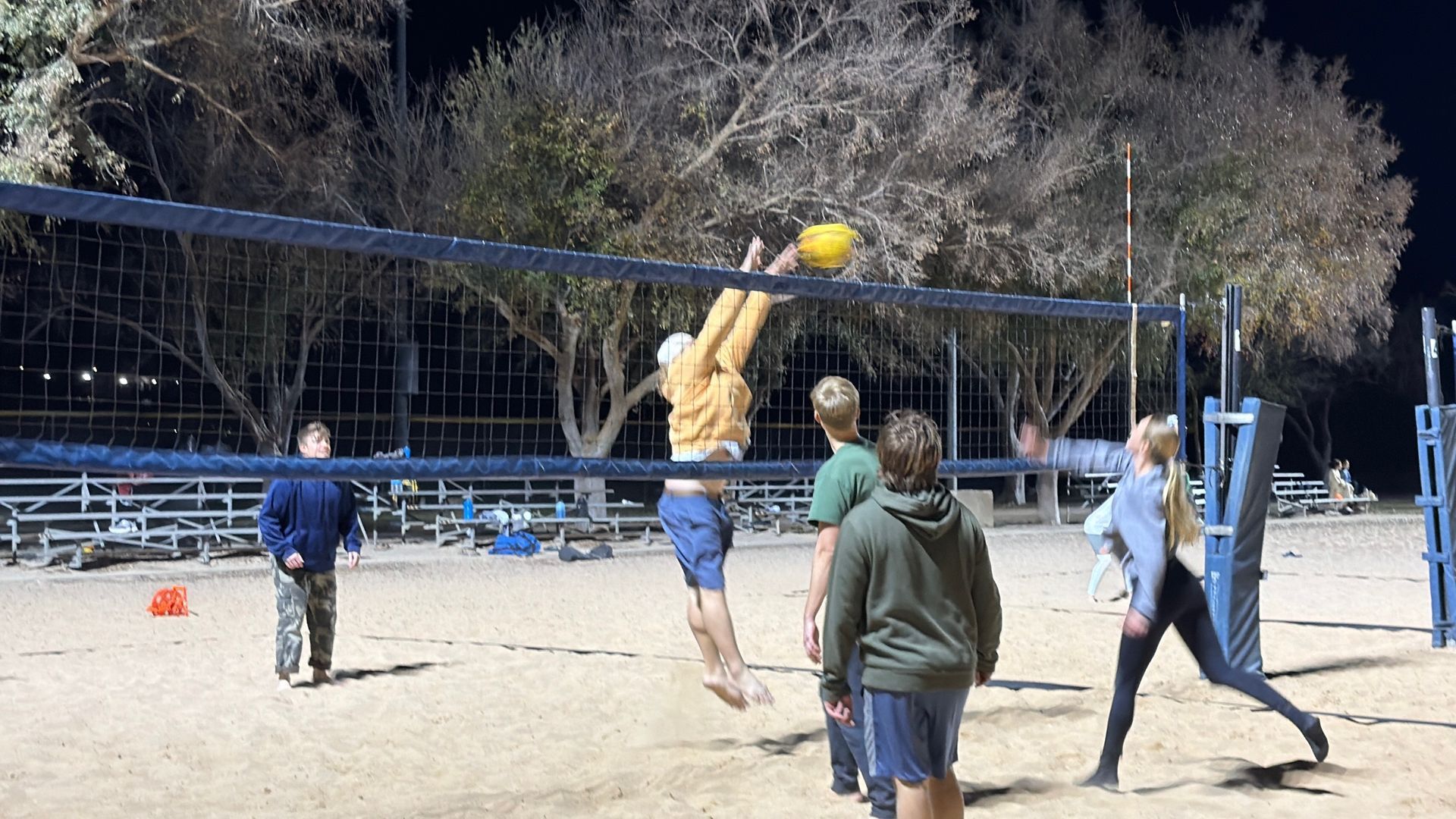 People playing volleyball at night on a sandy outdoor court, one jumping to hit the ball.